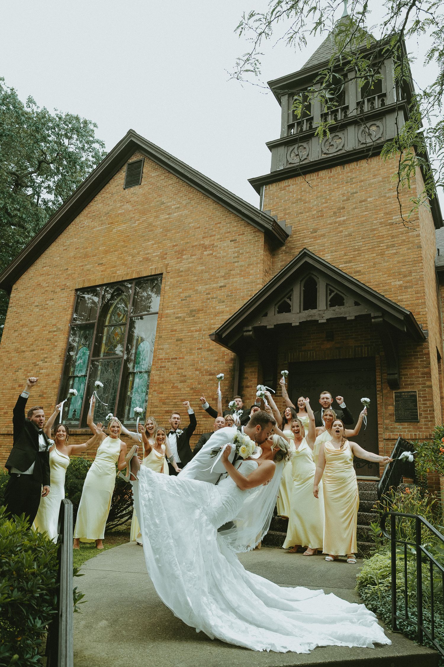 Bride and groom share a romantic dip kiss on church steps surrounded by cheering wedding party in yellow dresses.