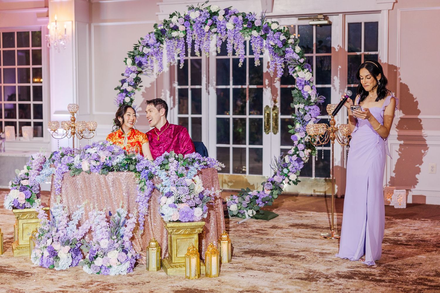 A woman in lavender gown speaks at a wedding reception with couple seated at floral-decorated sweetheart table.