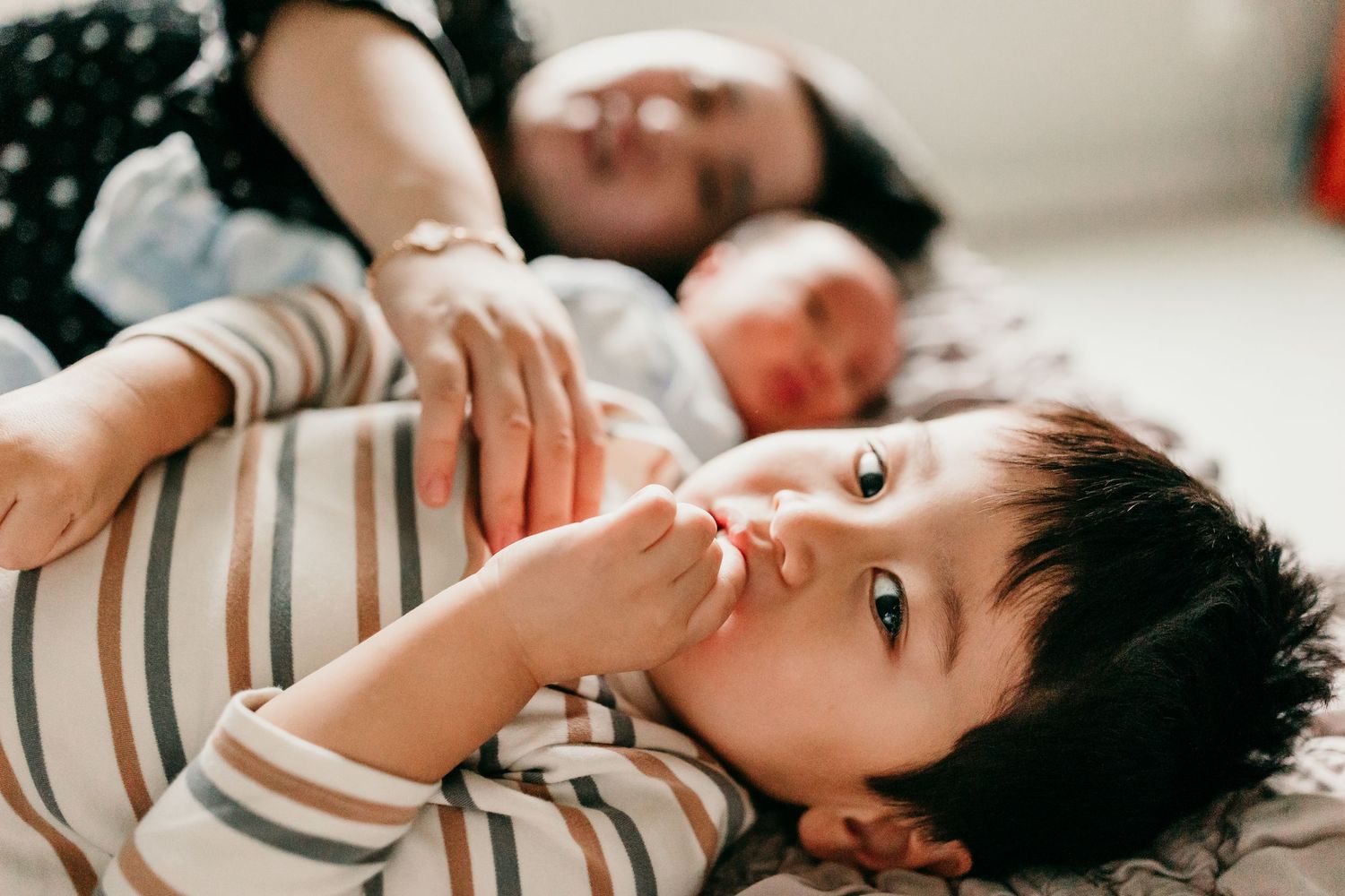 Close-up of curious Asian toddler boy lying down with parents and newborn baby in background.