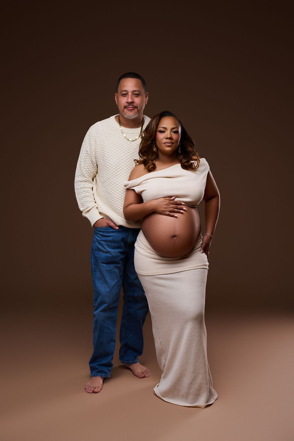 A couple poses for a maternity photo, man in cream sweater and jeans beside pregnant woman in elegant white dress. NJ Maternity Photography
