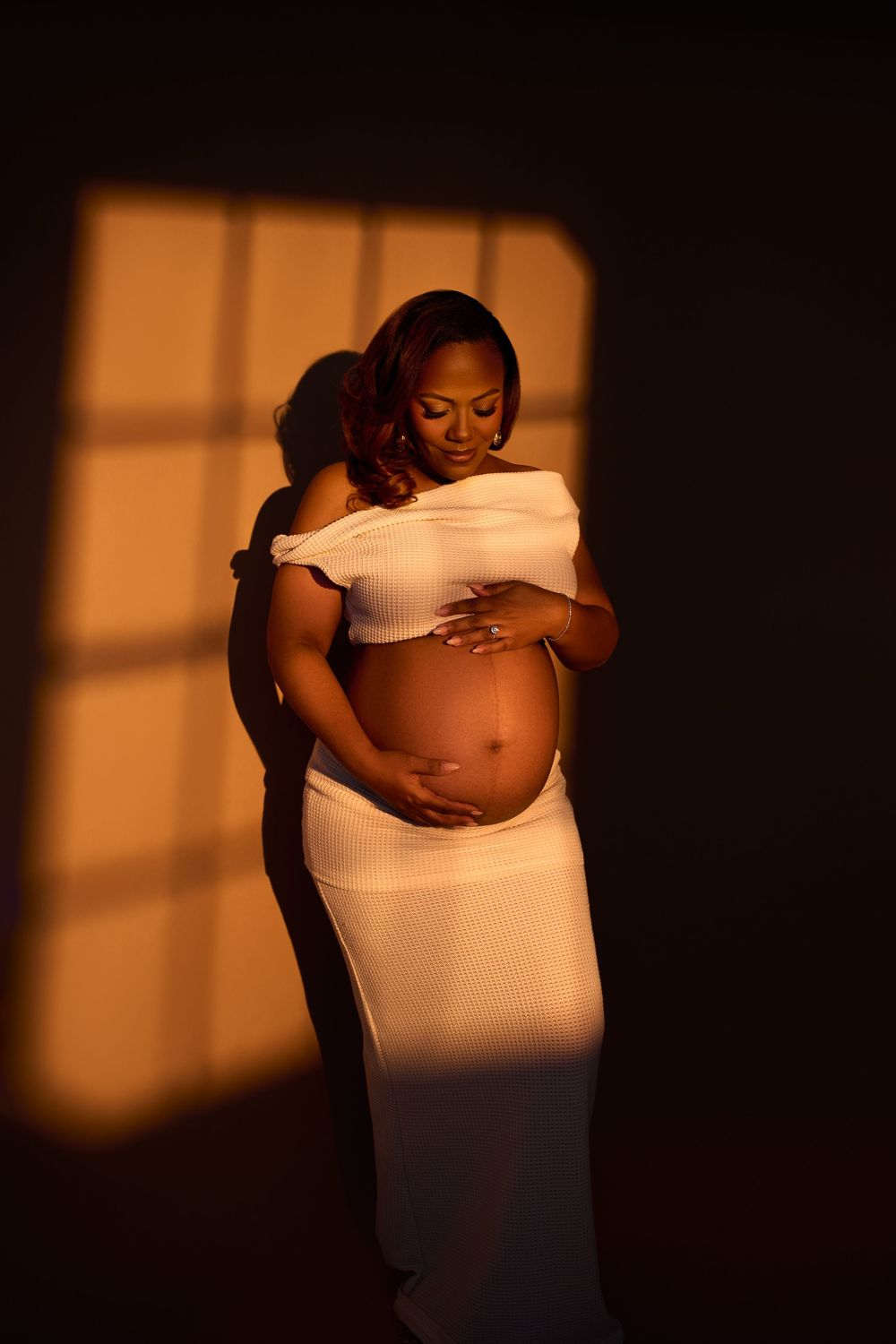 Pregnant woman in white off-shoulder dress cradling her baby bump, in warm golden window light against dark background. Gricel's photography