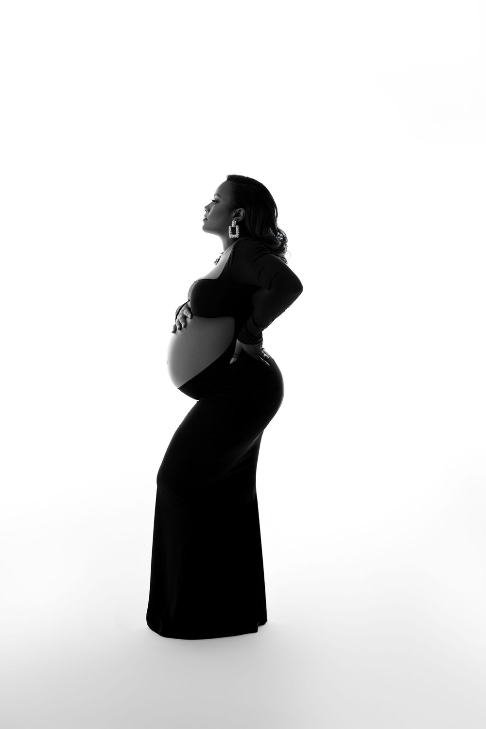 Elegant pregnant woman in black dress poses in silhouette against white background, cradling baby bump. NJ Maternity Photography