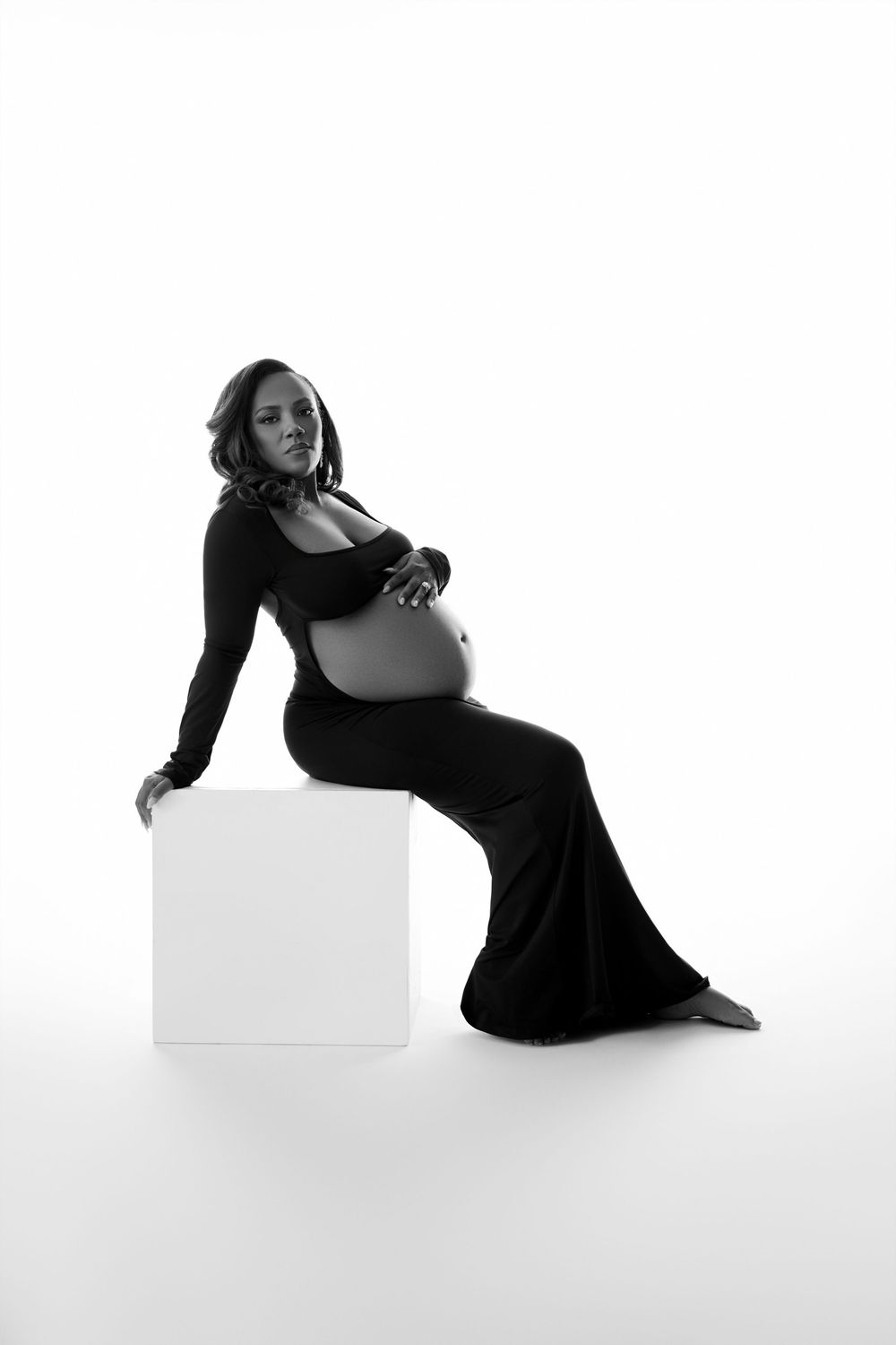 Black and white maternity photo of a pregnant woman in a long dress leaning on a white cube, cradling her belly. NJ Maternity Photography