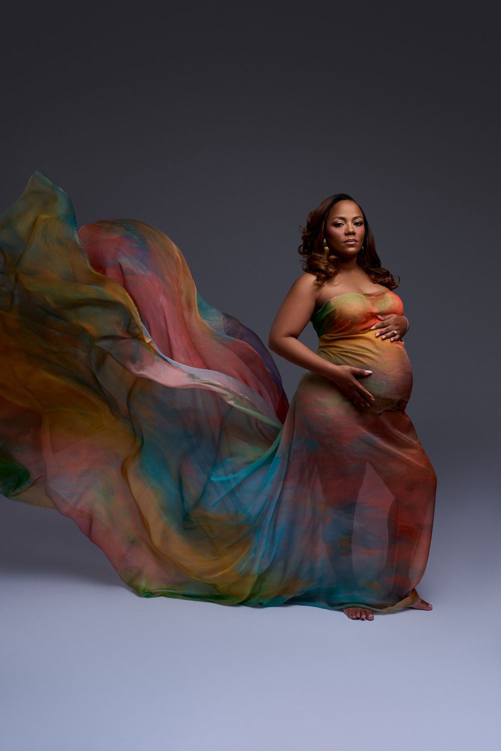 Pregnant woman in flowing multicolored sheer fabric gown billowing dramatically against gray studio background.