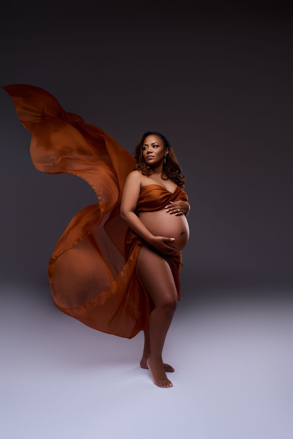 Pregnant woman draped in flowing rust-orange fabric against dark backdrop, cradling baby bump in elegant maternity photo.