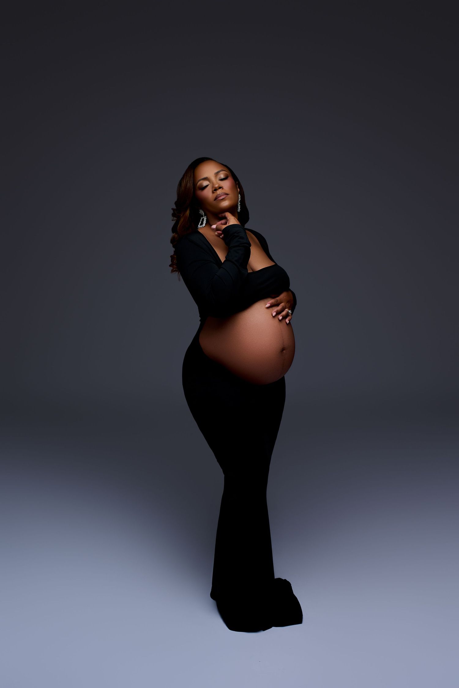 Pregnant Black woman in elegant black gown cradles bare baby bump in dramatic studio portrait. NJ Maternity Photography Gricels Photography