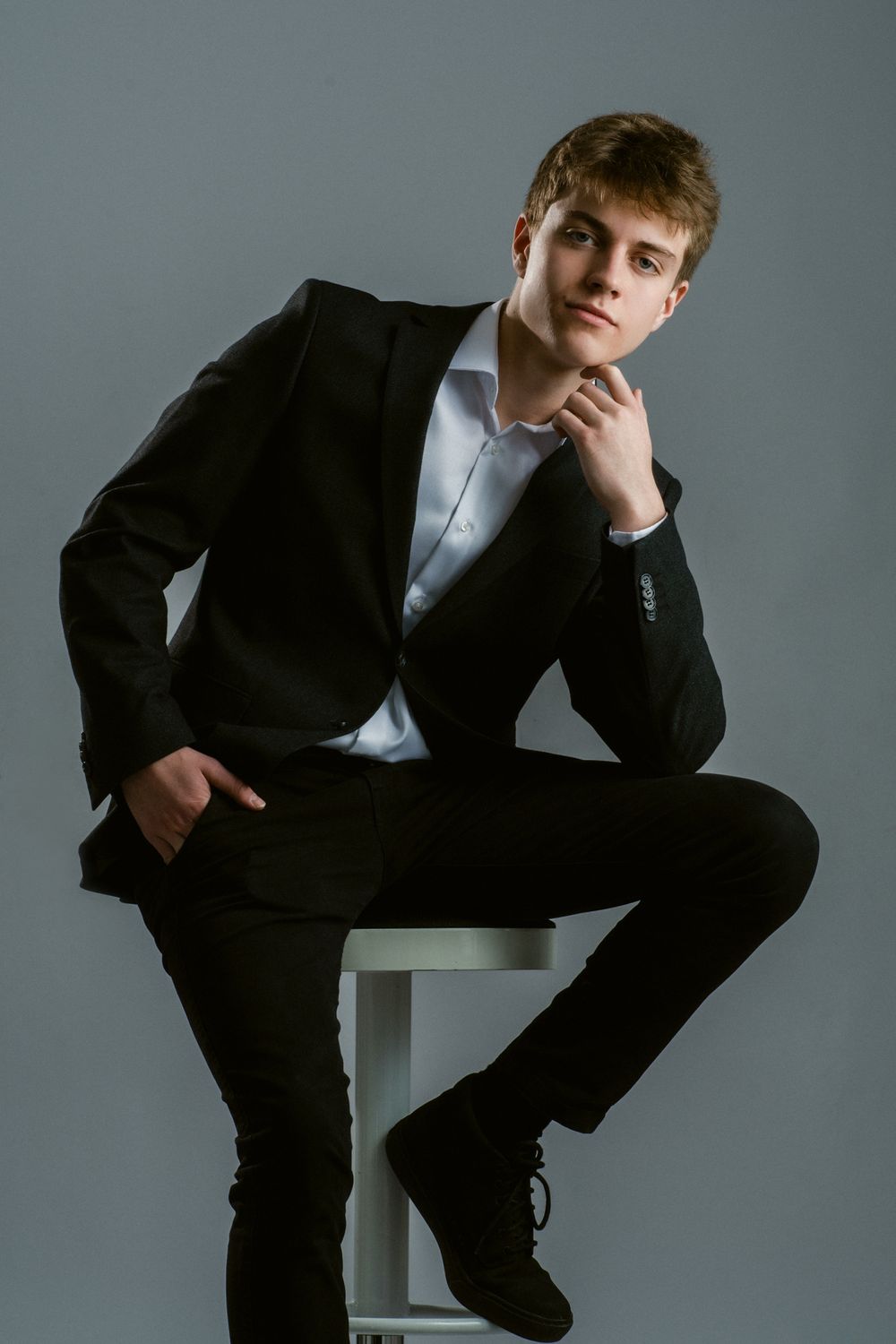 Professional studio portrait of young man in black suit sitting on stool against gray background.