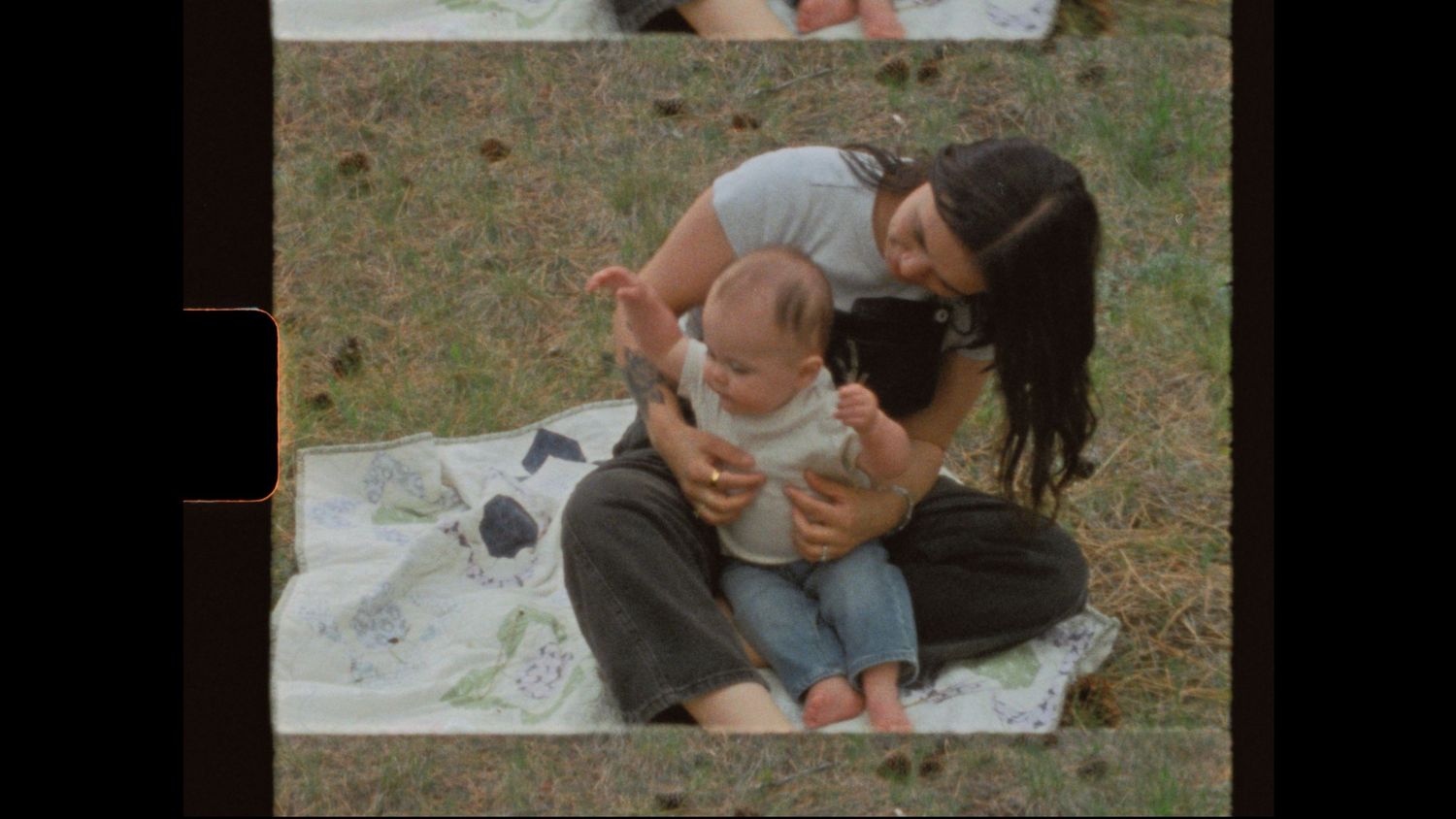 A mother sits on a blanket outdoors, lovingly holding her baby on her lap in a sunny park setting.