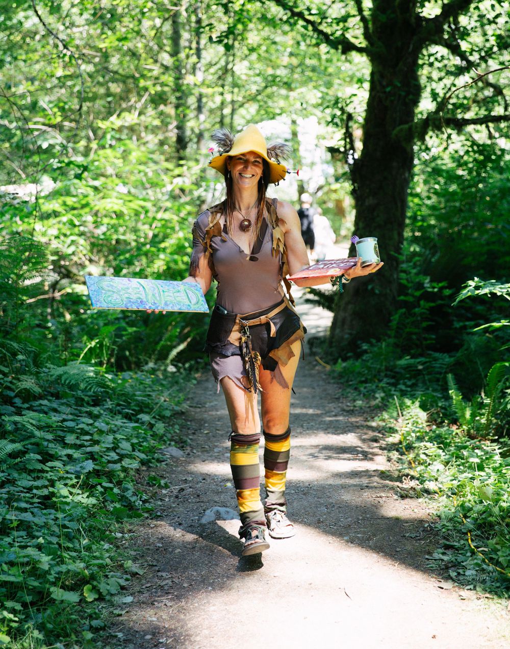 Woman in explorer costume with yellow hat holds map on forest trail in bright sunlight.