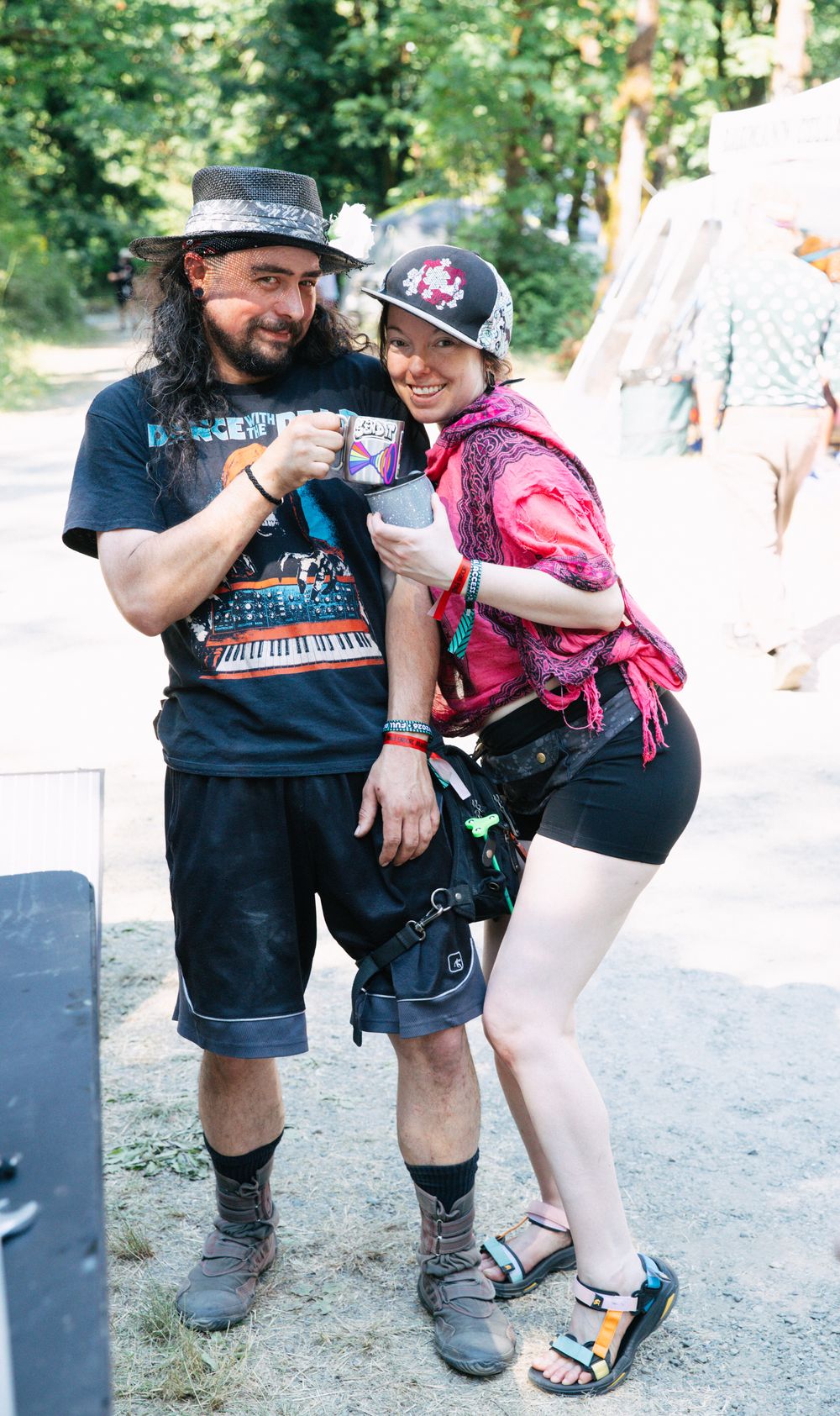 Two festival-goers in casual summer outfits toast mugs together outdoors at a music festival.