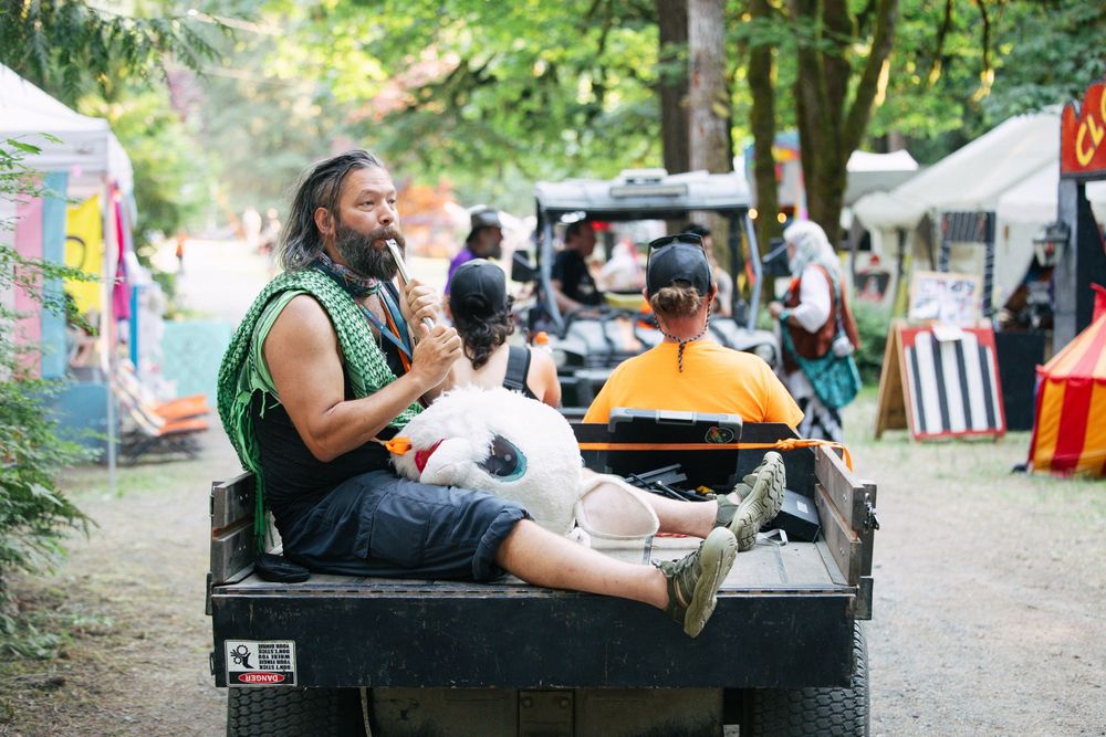 Man with dreadlocks holding a dog rides on the back of a flatbed truck at an outdoor festival.