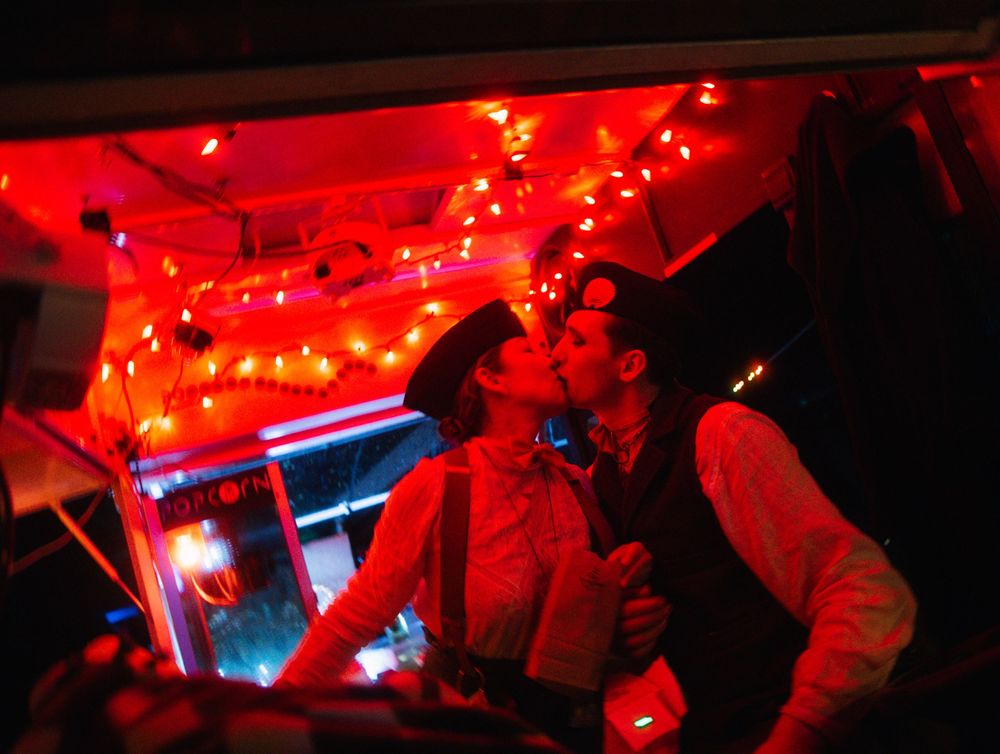 Couple kissing under red string lights in a dimly lit, festive venue with warm holiday ambiance.