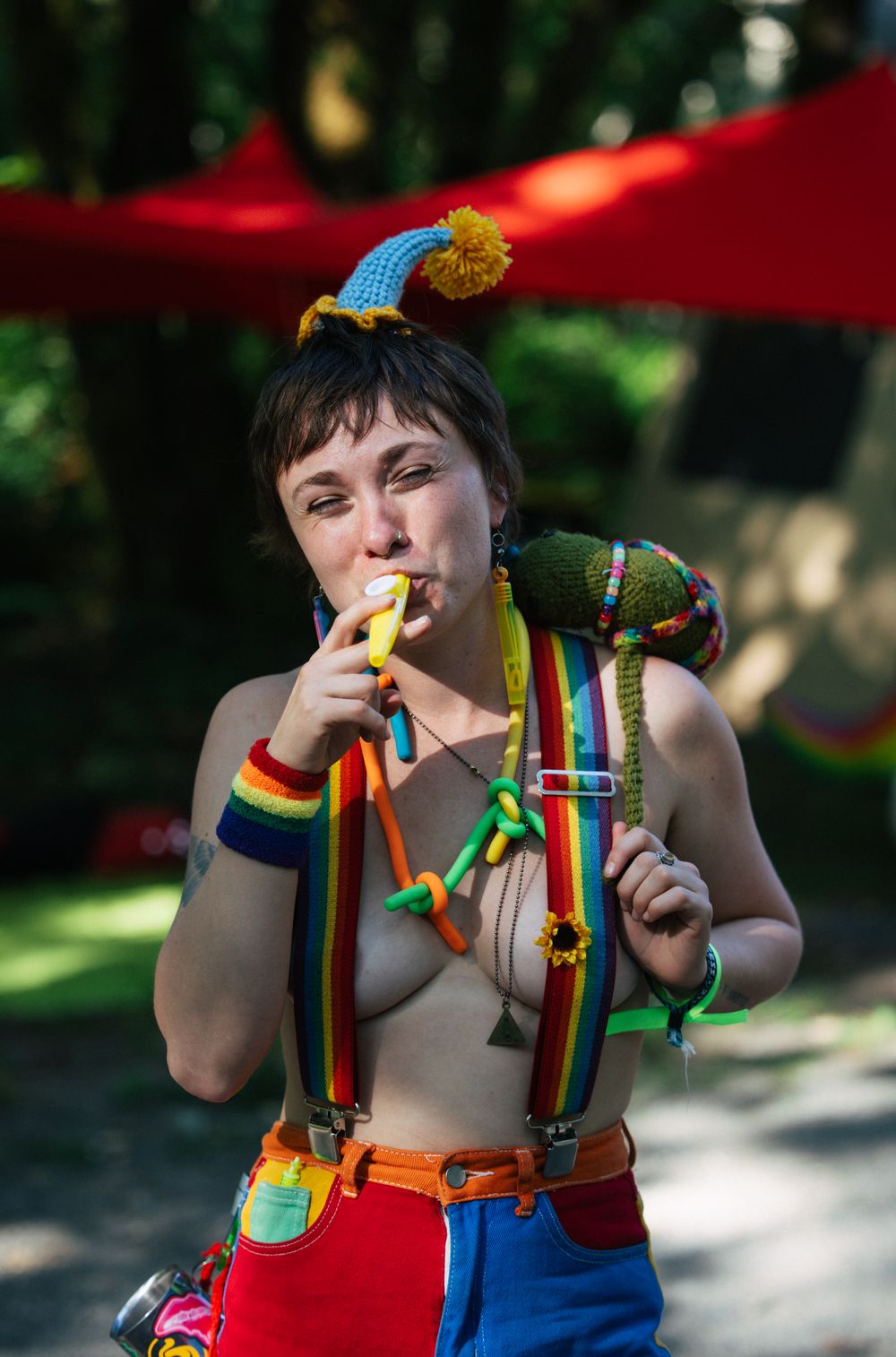 A woman in colorful rainbow suspenders and a crocheted hat plays a small instrument outdoors at a festive event.