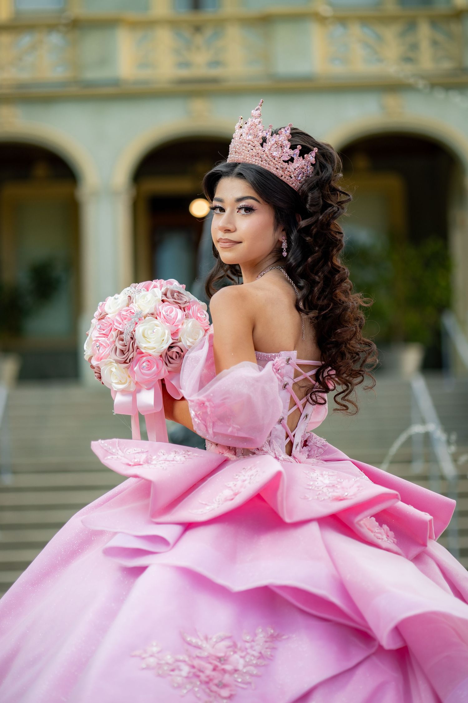 Young woman in pink quinceañera ball gown and crown holds a bouquet on decorative steps.