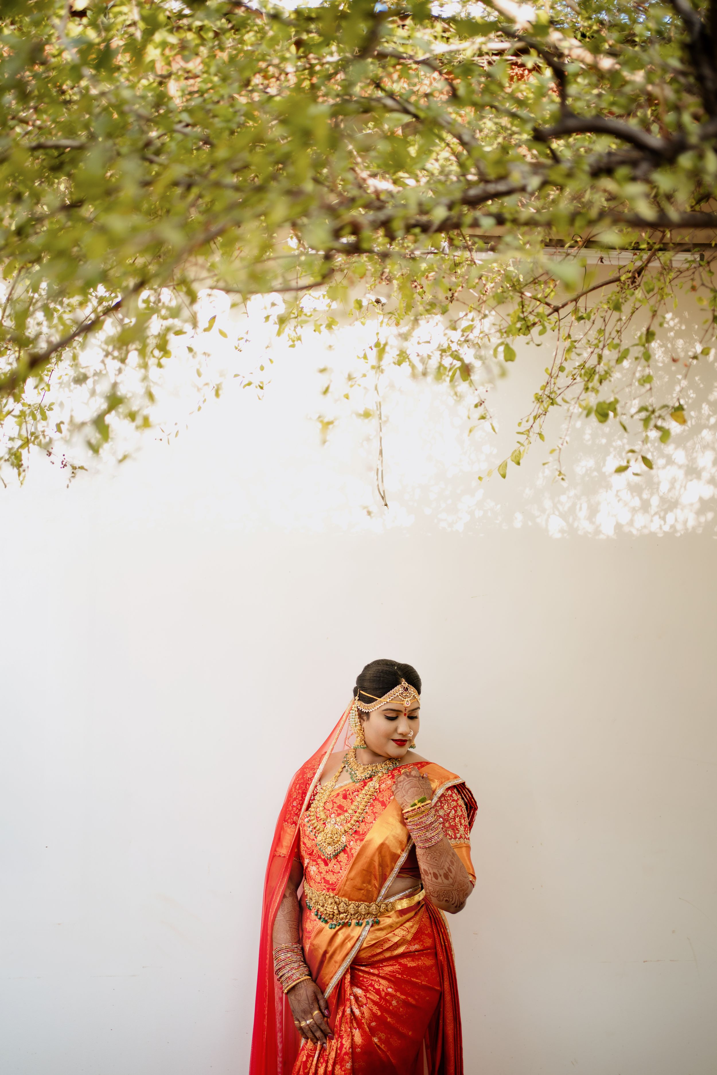 Woman in red and white saree with henna hands poses gracefully in dim light against dark curtains.