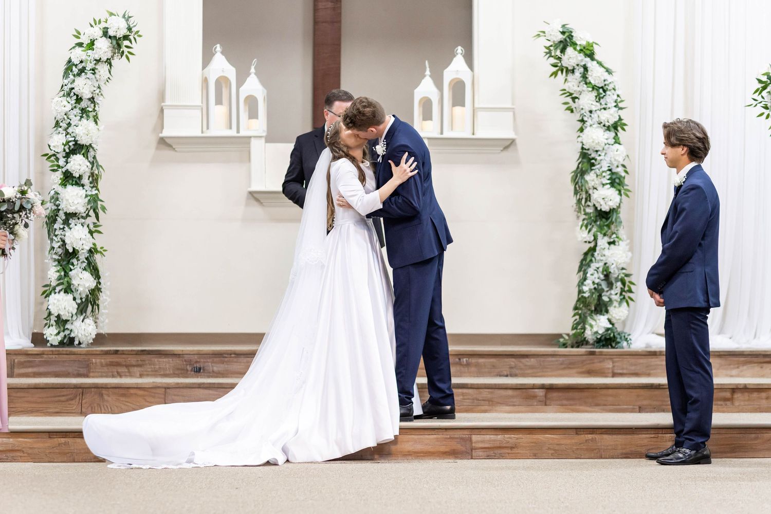 Bride and groom share first kiss at altar during elegant white church wedding ceremony with floral arrangements.