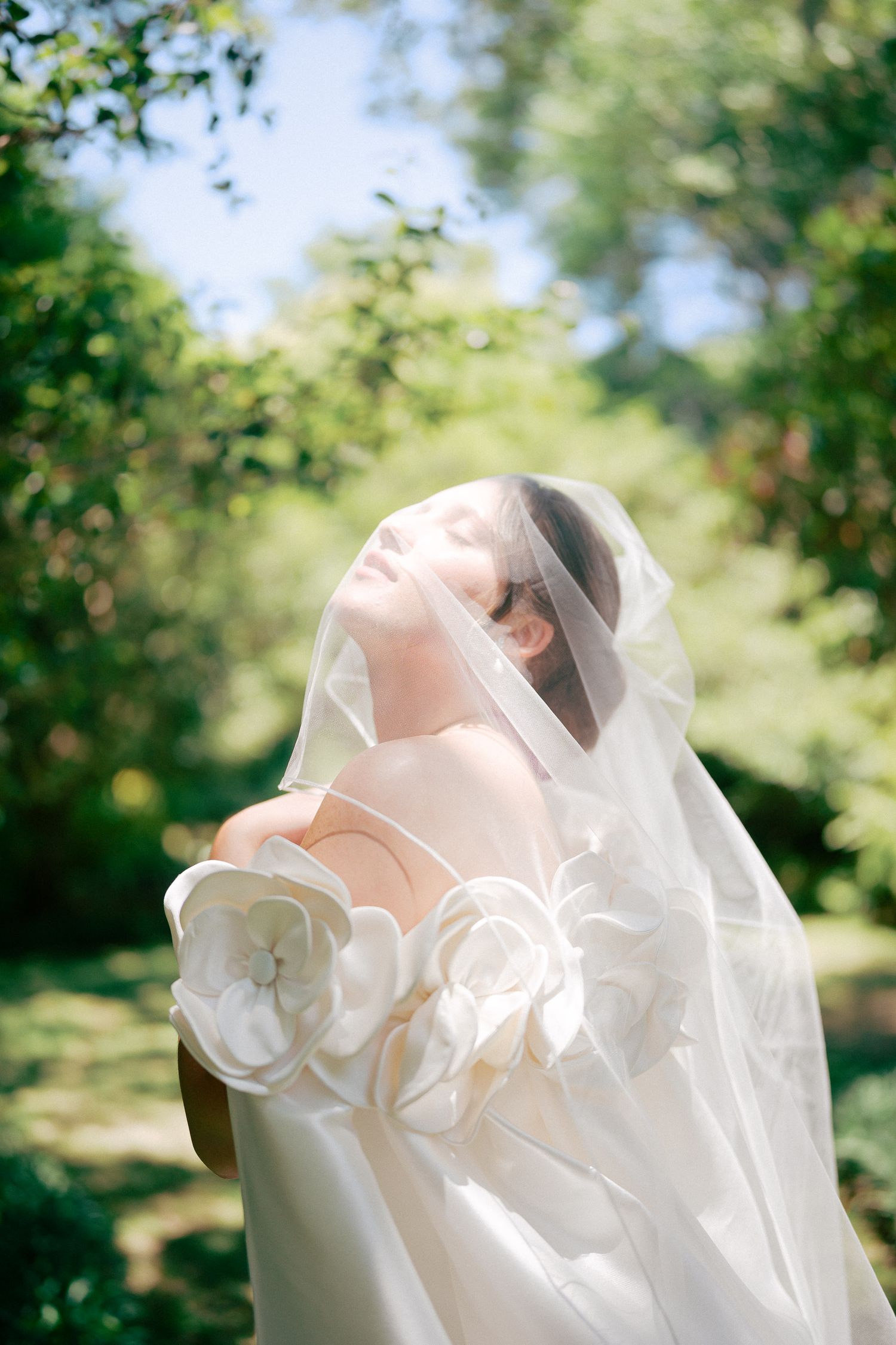 Bride in white floral gown and veil tilts head back in sunlit garden.