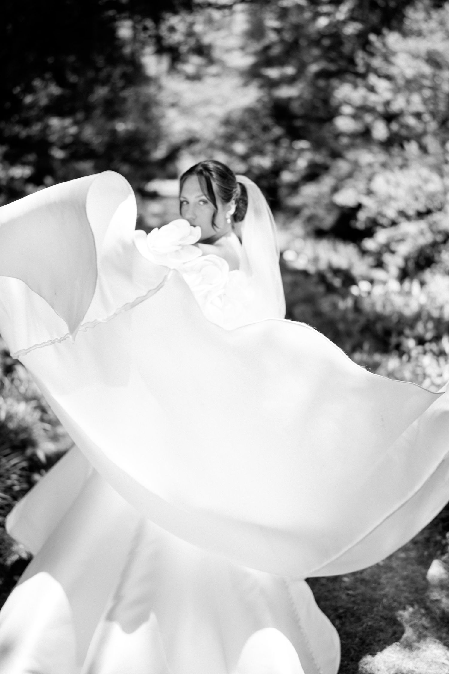 Black and white photo of a bride twirling her flowing wedding gown outdoors among lush green trees.