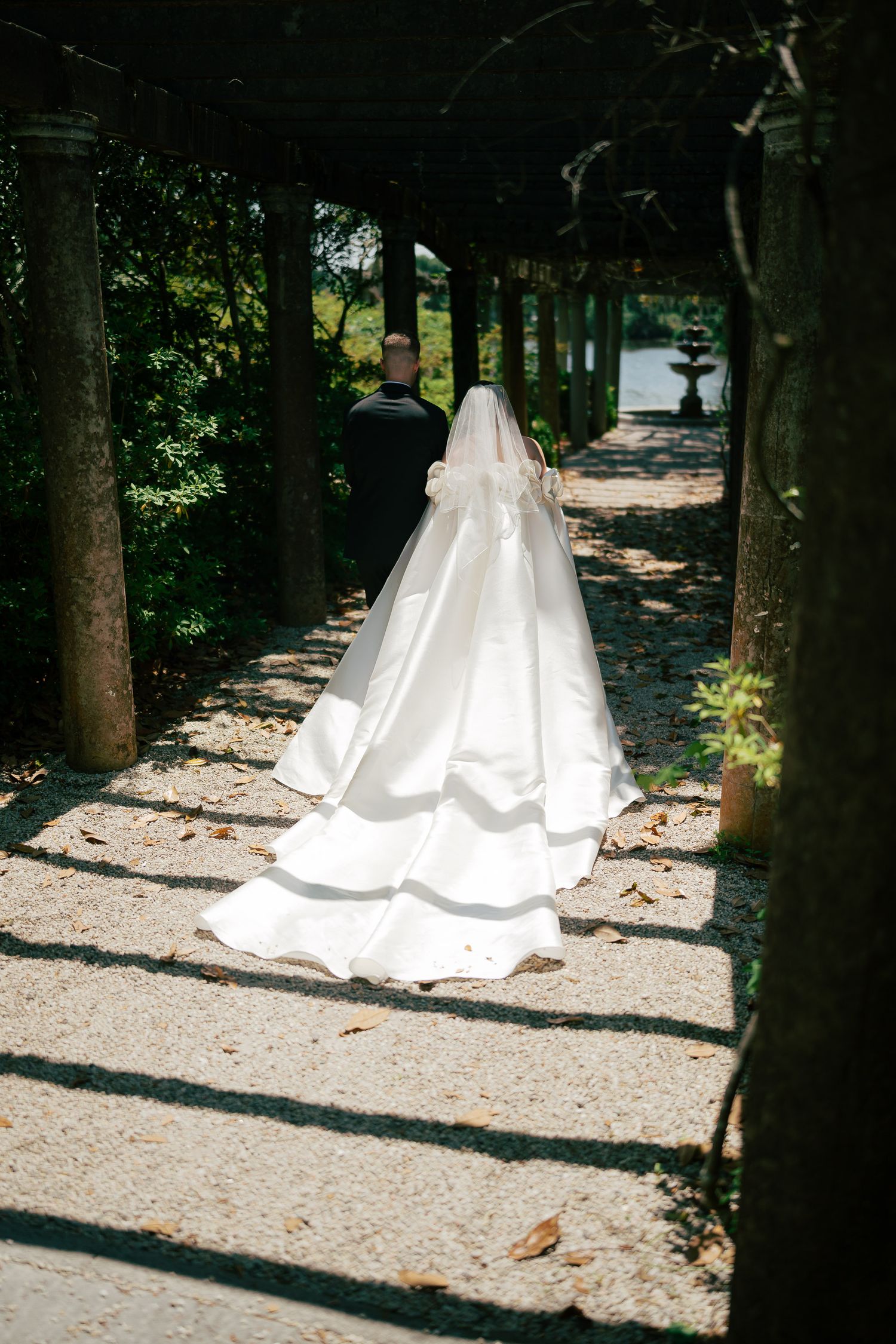 Bride in elegant white gown with long train walks through a sunlit garden colonnade toward groom.
