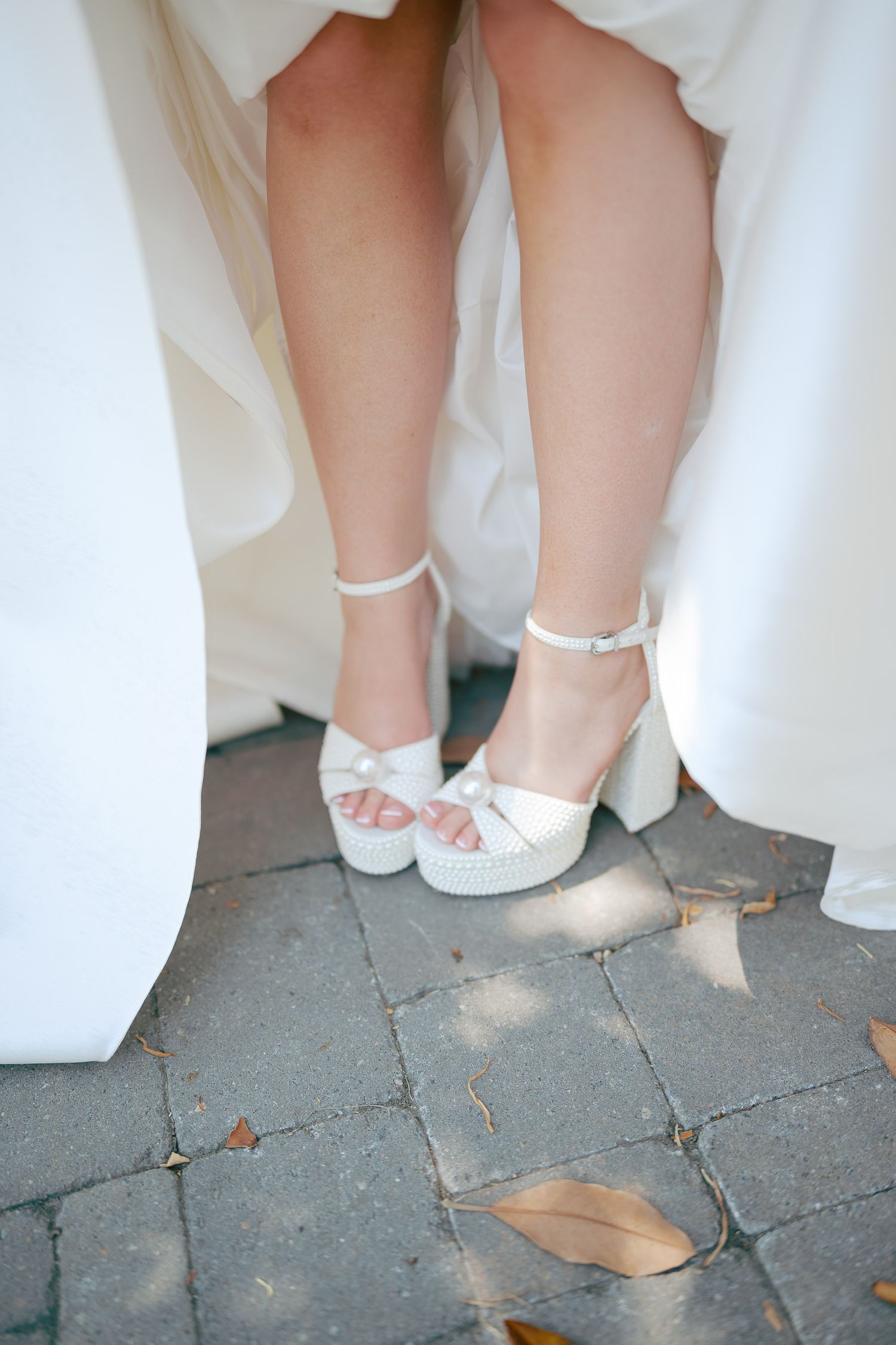 Bride wearing white platform heeled sandals with ankle straps and bow detail on stone pavement.