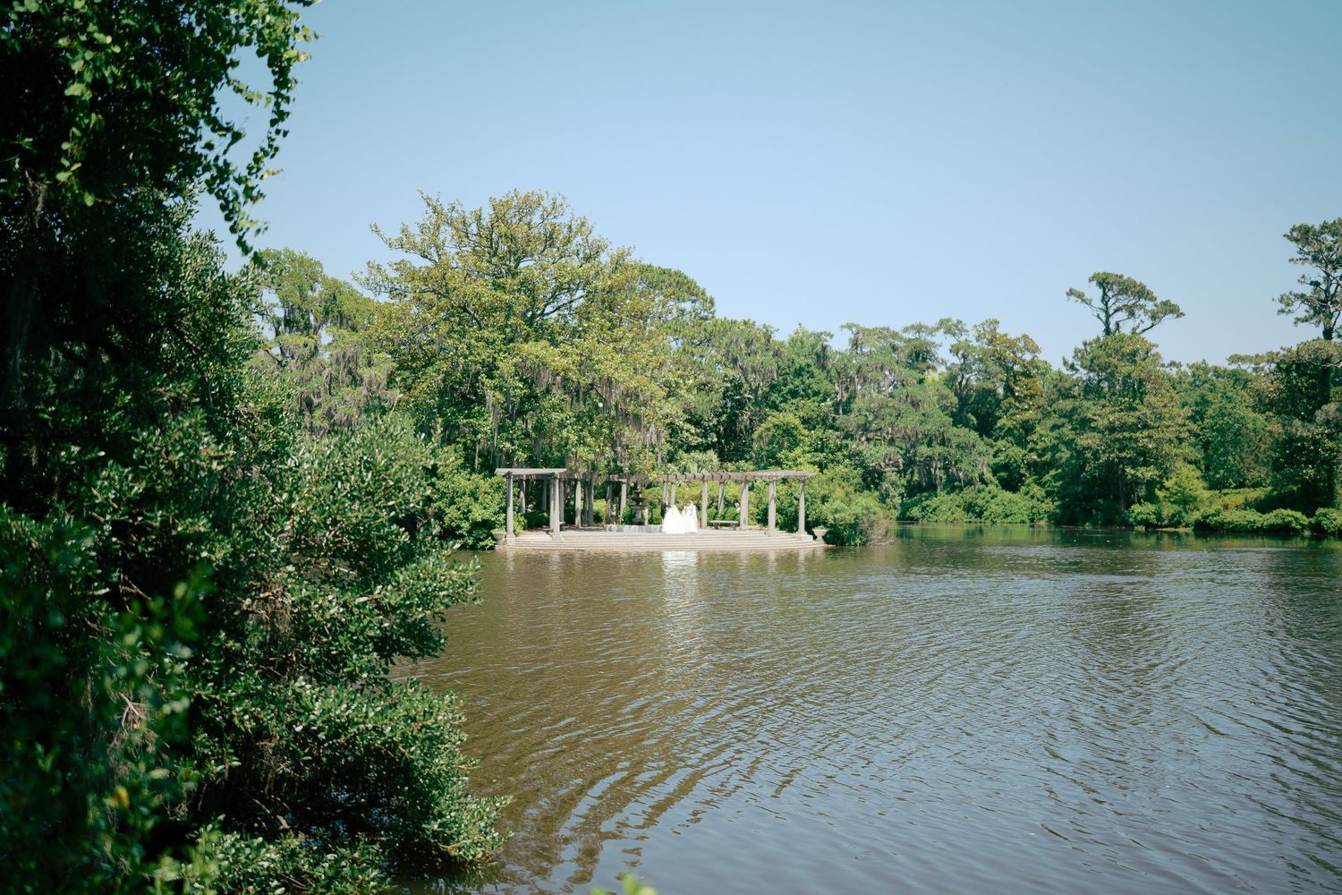 Serene lake surrounded by lush green trees with a white gazebo and dock visible in the background on a clear day.
