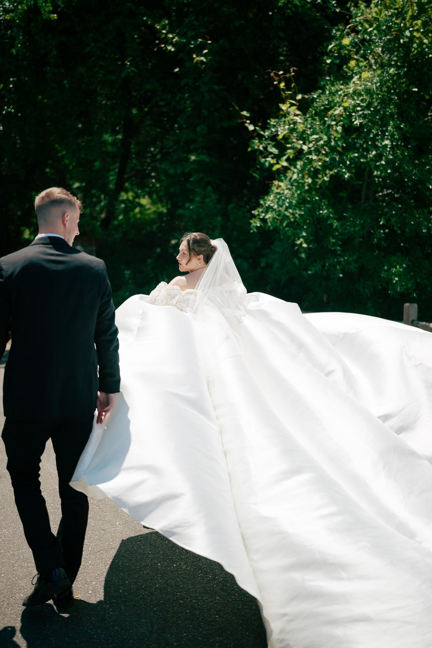 Bride in flowing white gown with long train walks with groom in black tuxedo through lush green park.