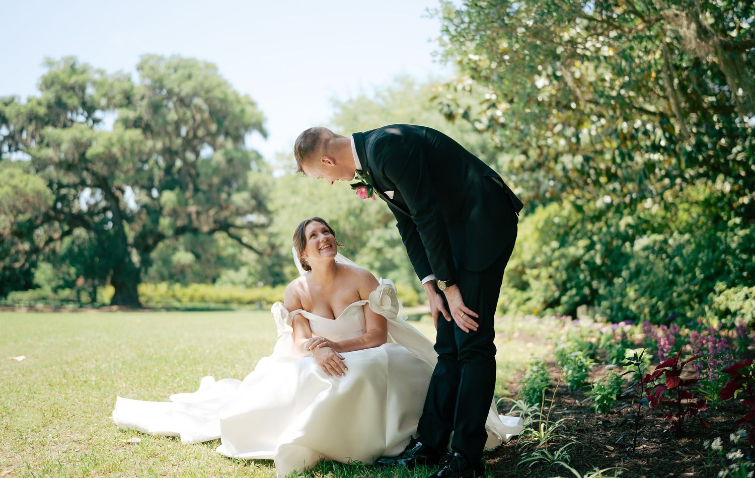 Bride in white off-shoulder gown sits on grass as groom in black tux leans toward her in lush garden setting.