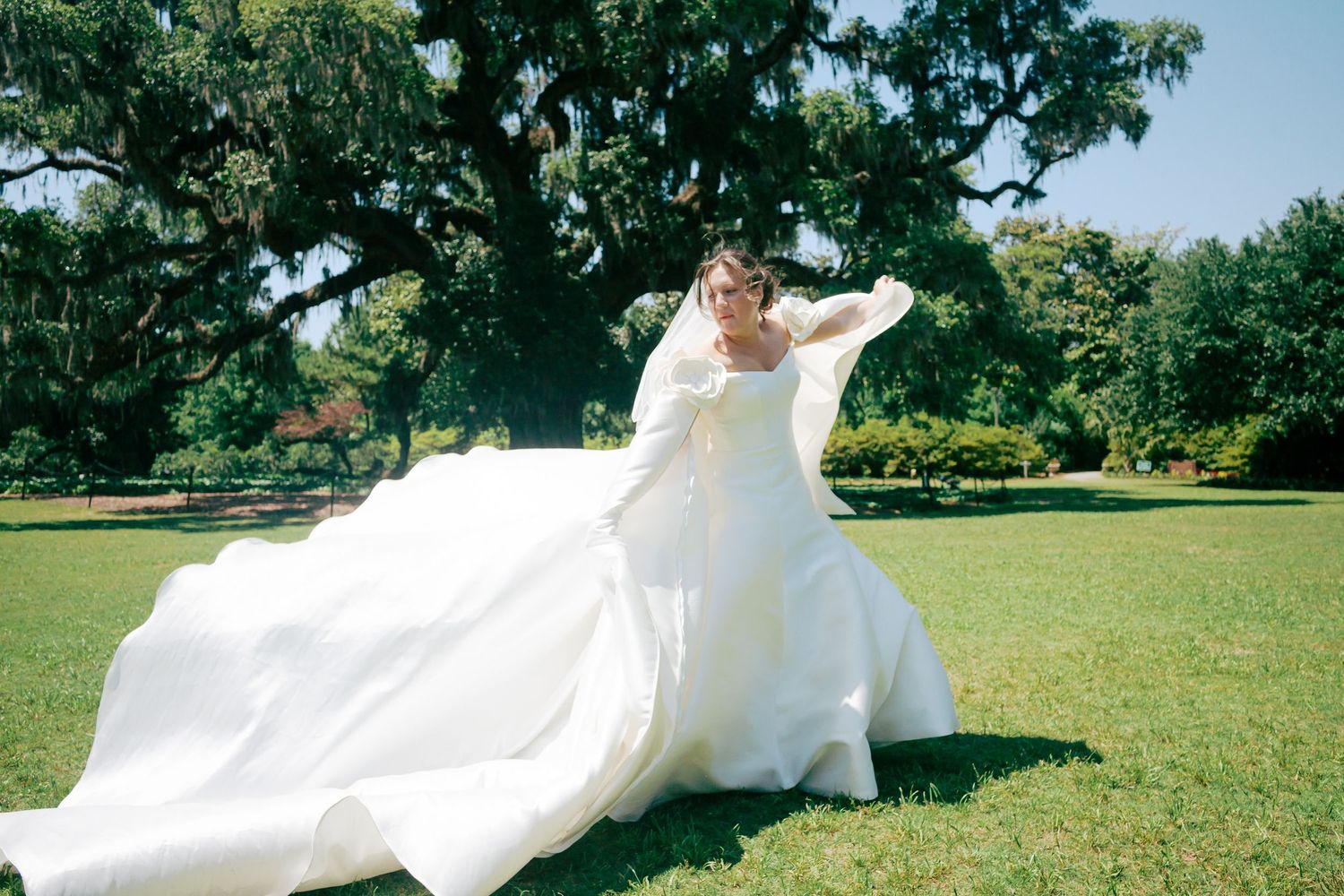 Bride in flowing white wedding gown twirling on green lawn with large oak tree backdrop on sunny day.