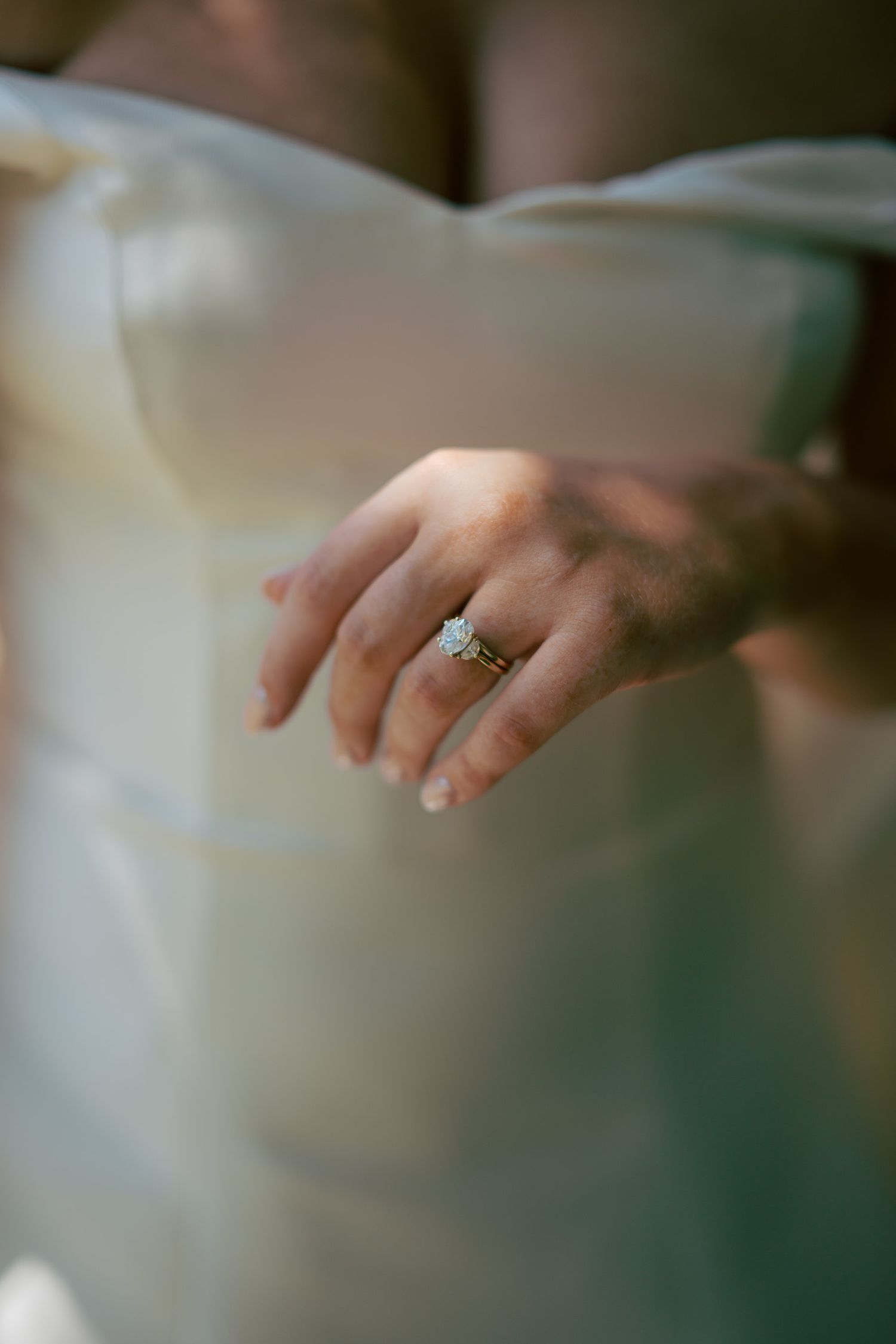 Close-up of a bride's hand adorned with a diamond engagement ring and gold wedding band against a white bridal gown.