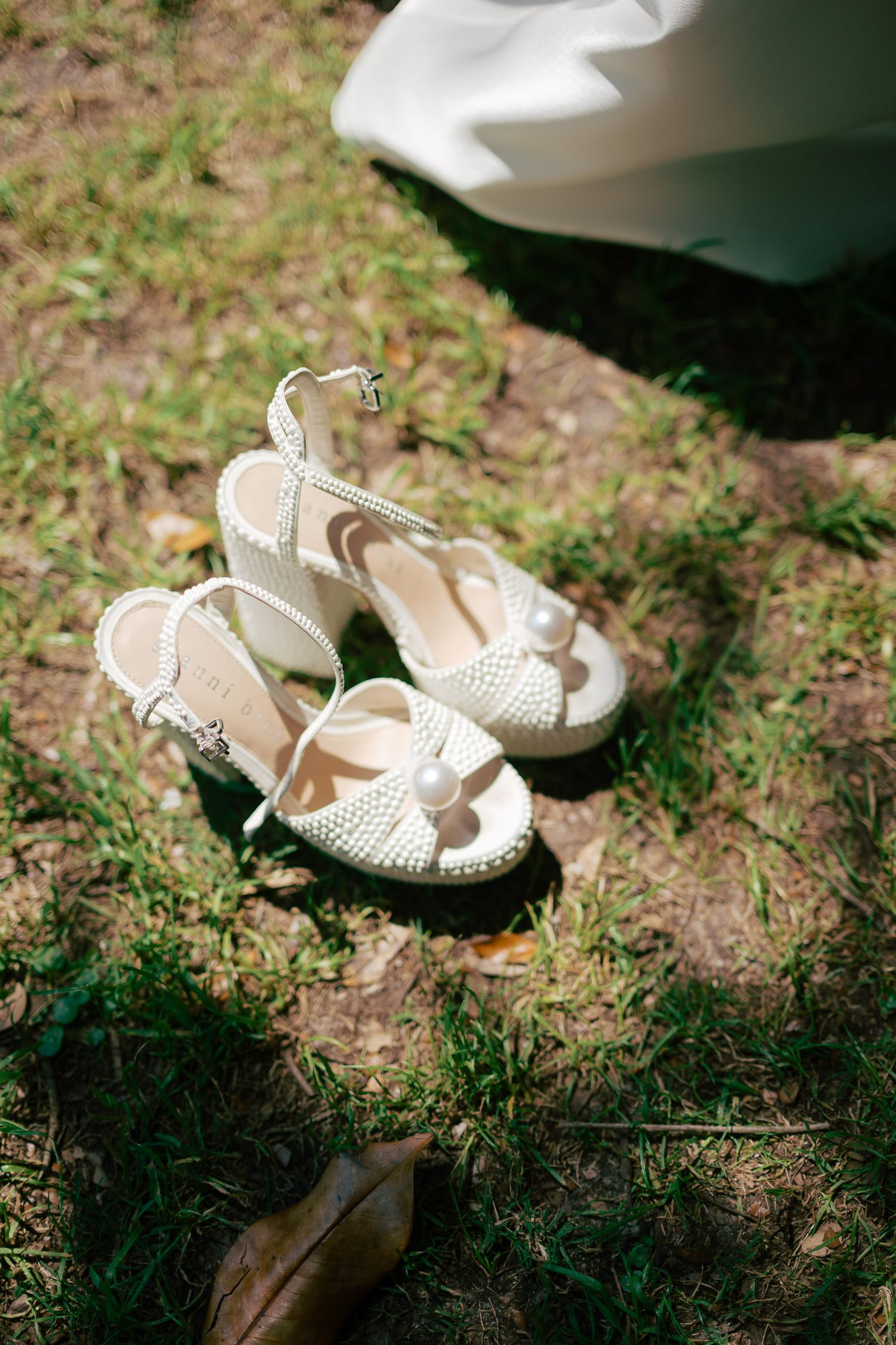 White pearl-embellished bridal heeled sandals placed on green grass outdoors on a sunny wedding day.
