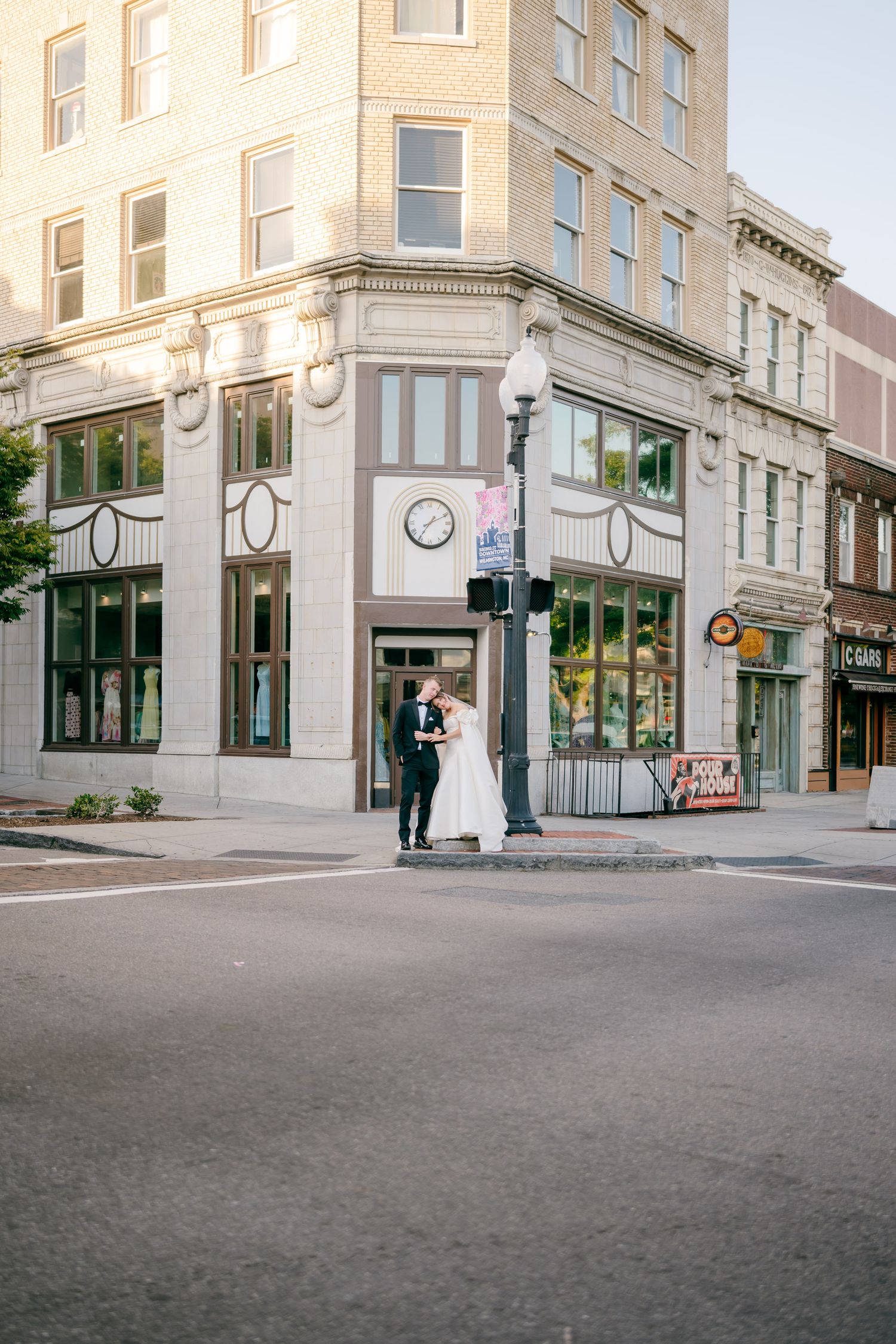 Wedding couple shares romantic kiss at urban street corner near classic building with clock at golden hour.