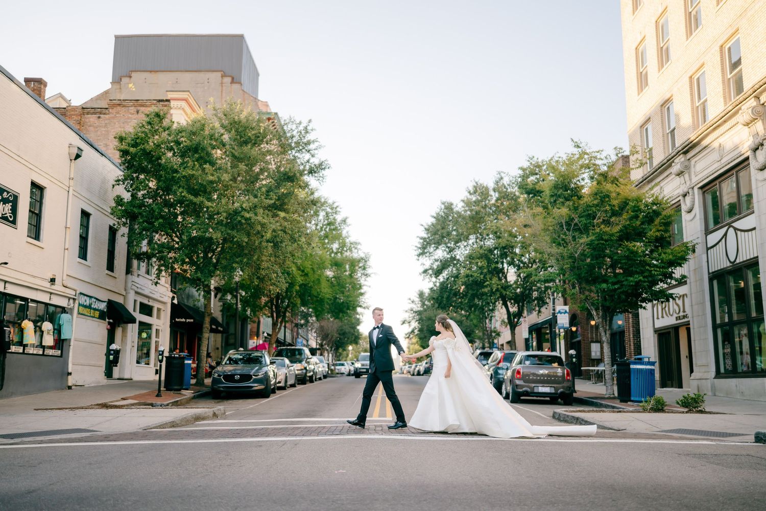 Bride and groom crossing a charming urban street in wedding attire, with tree-lined buildings in the background.