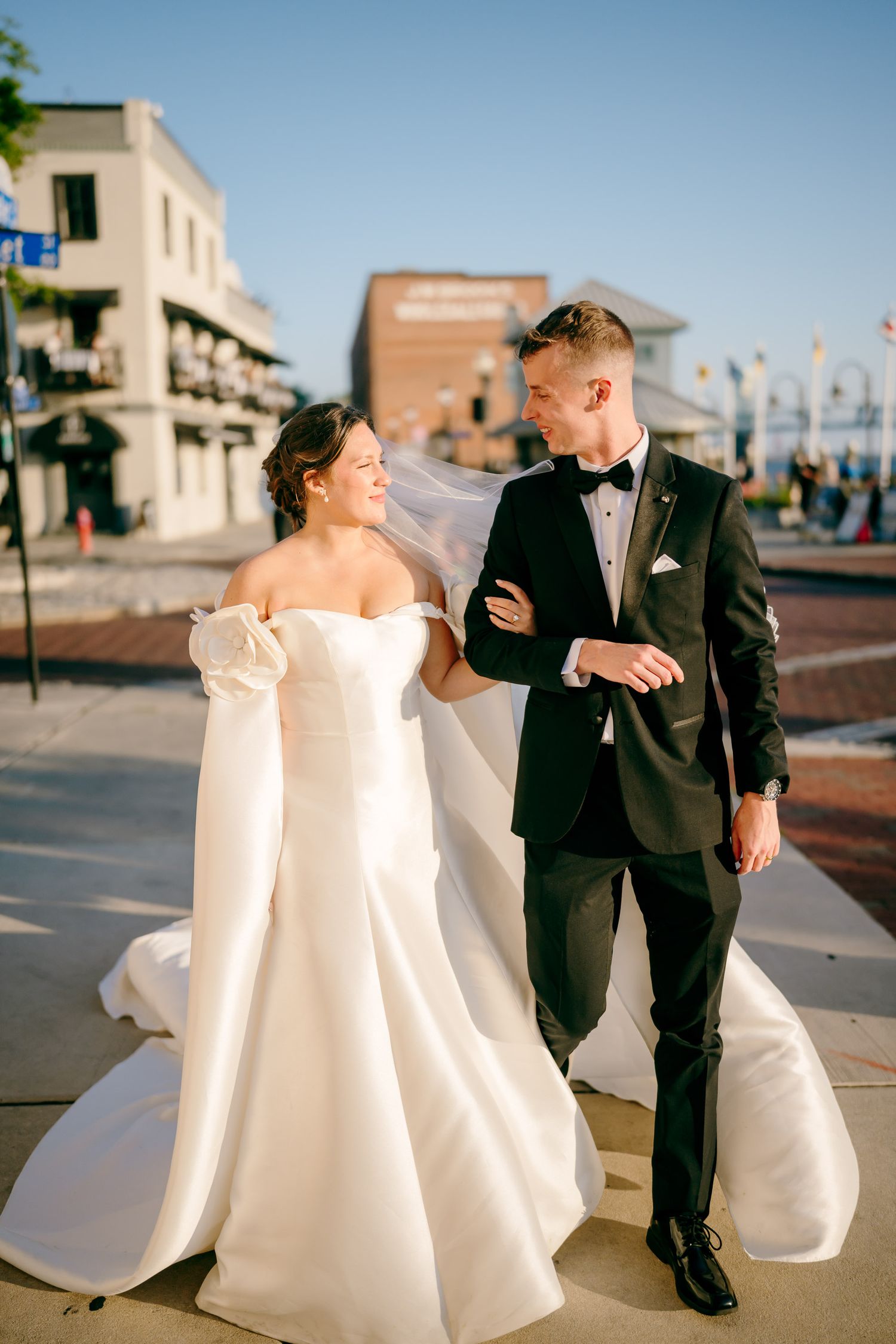 A bride in a white off-shoulder gown and groom in black tuxedo walk together outdoors on a sunny wedding day.