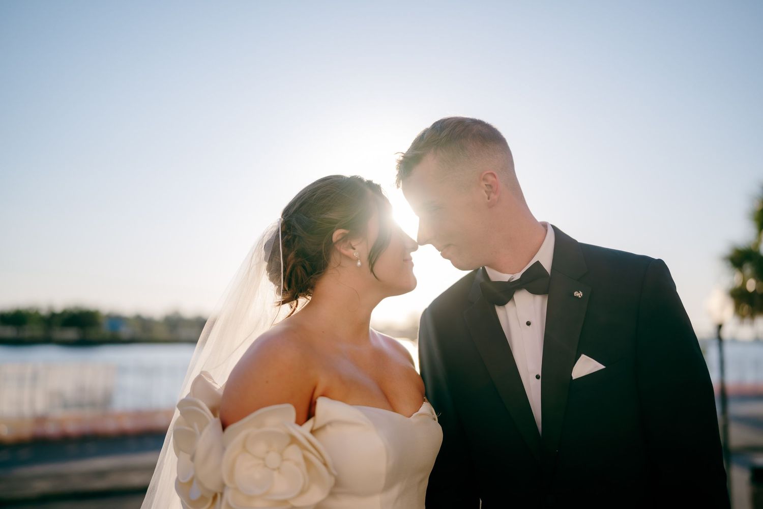 Bride and groom share romantic moment outdoors at sunset near waterfront, bride in strapless gown with veil, groom in black tuxedo.