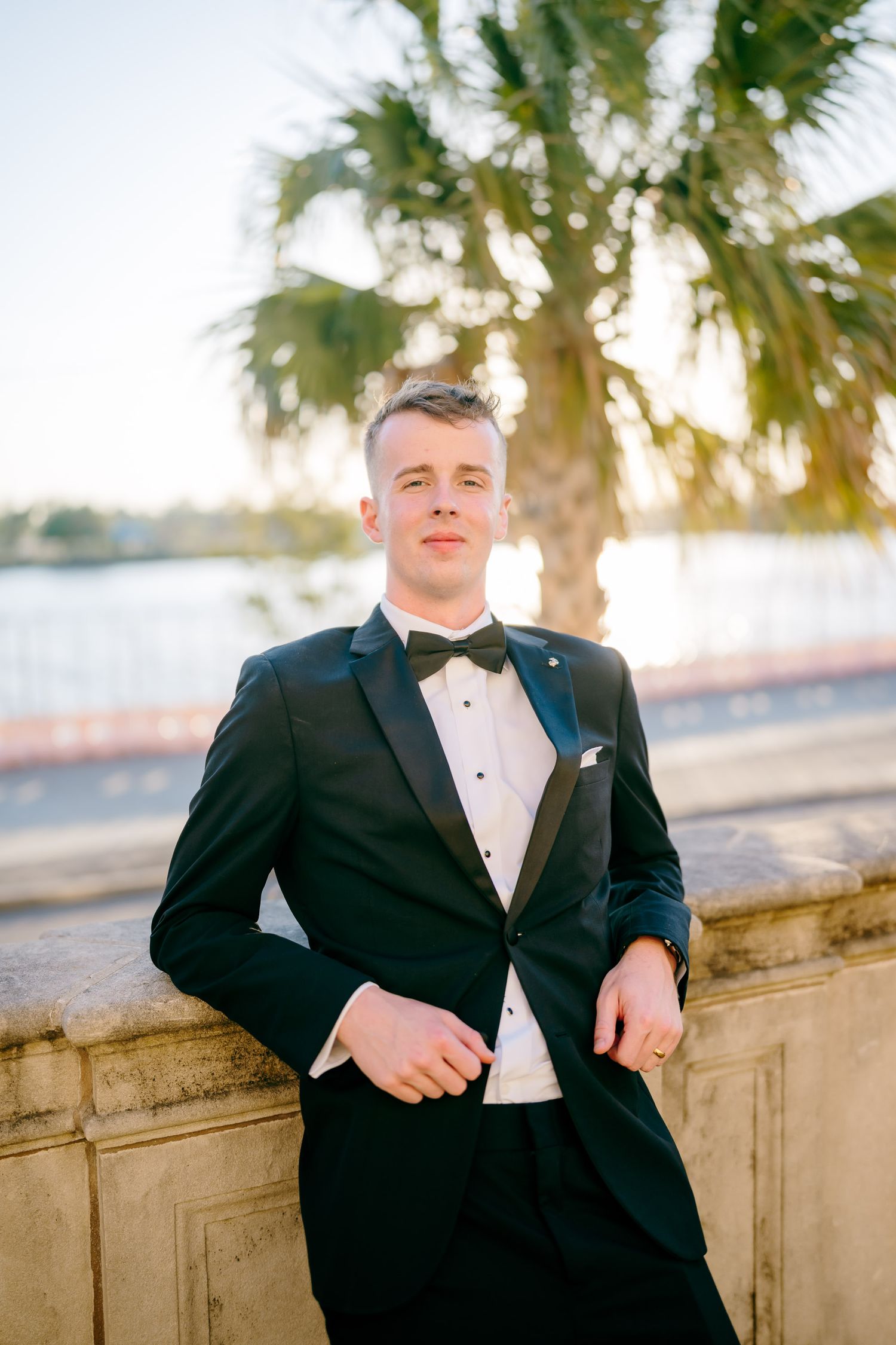 Elegant groom in black tuxedo with bow tie poses confidently near palm tree and waterfront at sunset.