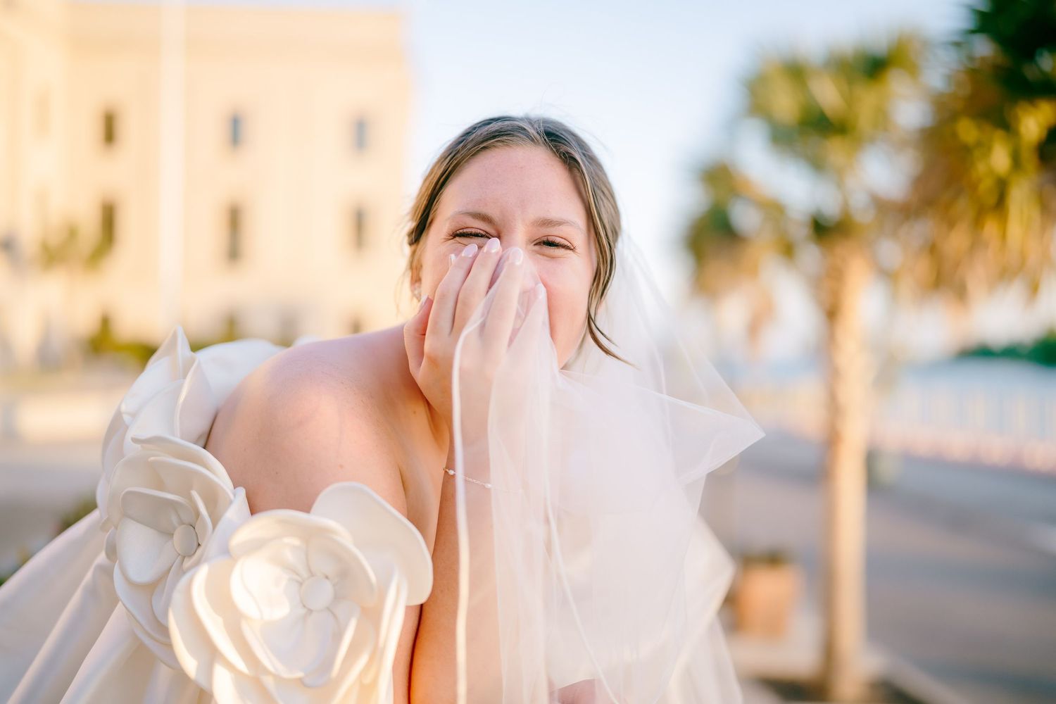 A joyful bride in a floral wedding gown laughs while holding her veil to her face outdoors.