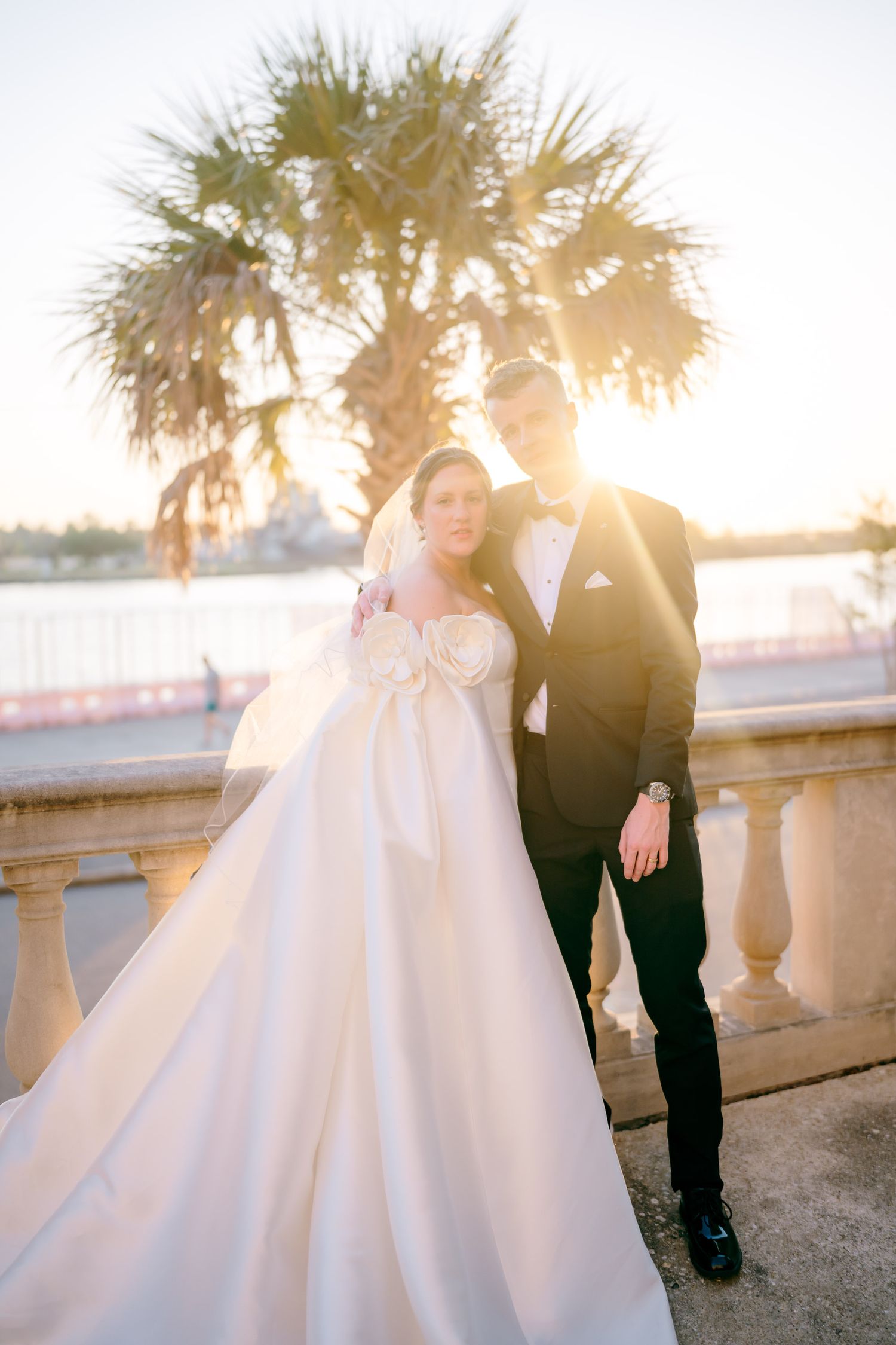 Bride in white ballgown and groom in black tuxedo pose on a sunlit terrace with palm trees and waterfront backdrop.