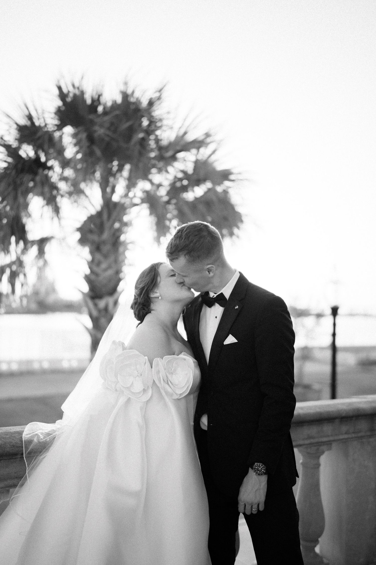 Black and white photo of a bride and groom sharing a romantic moment near a balustrade with a palm tree backdrop.