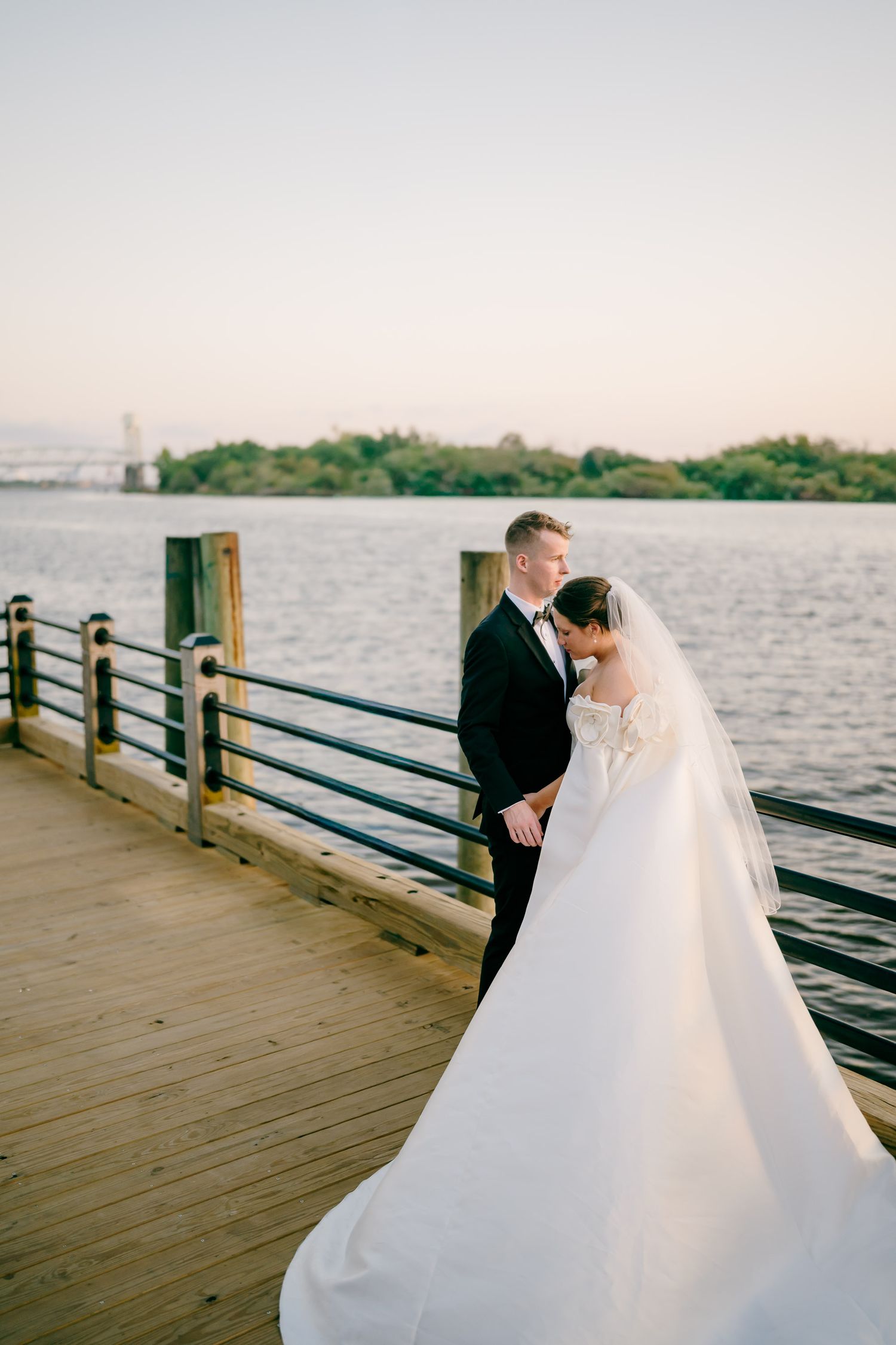 A bride and groom share a romantic moment on a wooden pier overlooking a serene waterway at sunset.