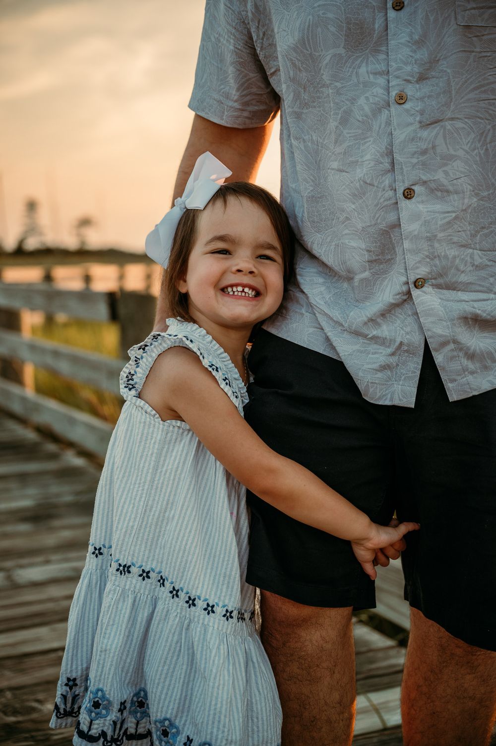 A smiling toddler girl in a white dress holds an adult's hand on a wooden pier at sunset.