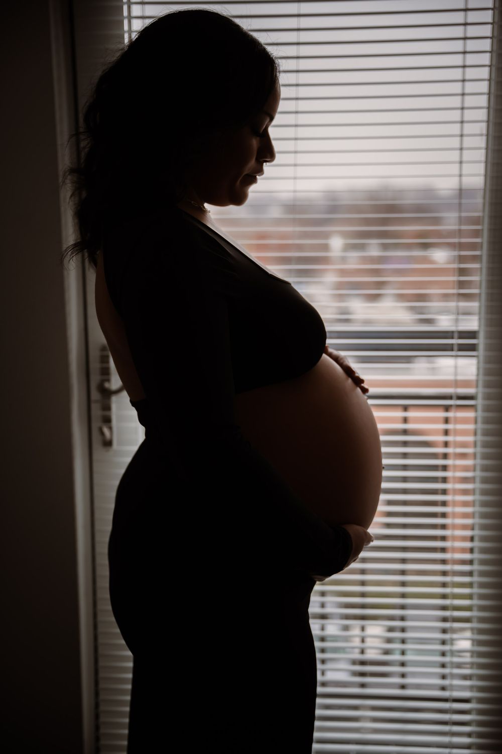 Silhouette of a pregnant woman standing by a window with blinds, cradling her baby bump in profile view.