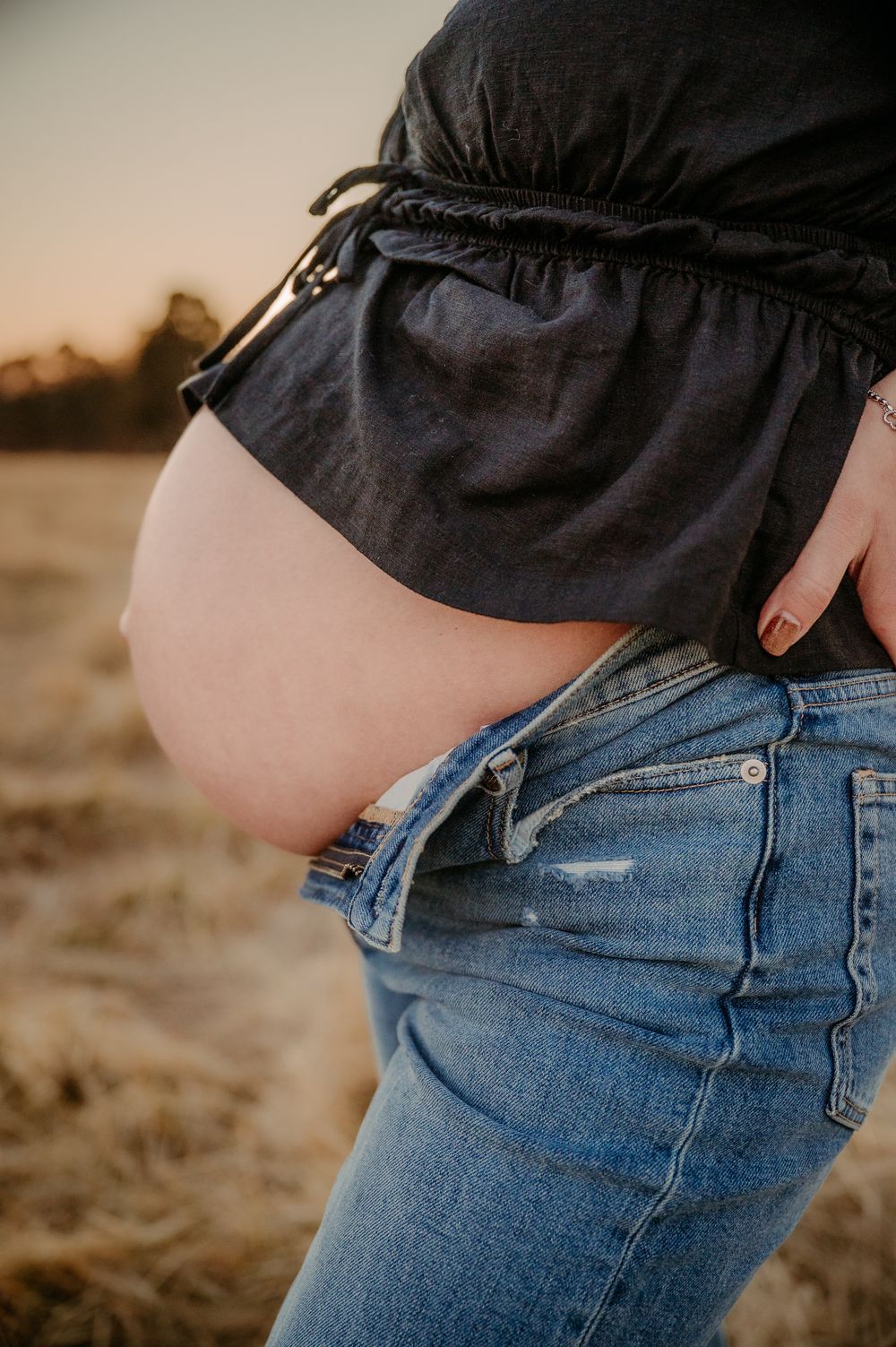 Close-up of pregnant woman's baby bump showing between black top and unbuttoned blue jeans at golden hour outdoors.