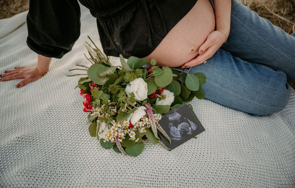 Pregnant woman sitting on blanket with floral bouquet and ultrasound photo, cradling baby bump outdoors.