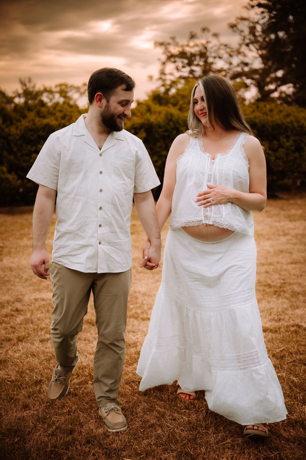 Pregnant couple holding hands walking outdoors in white outfits during golden hour maternity photoshoot.