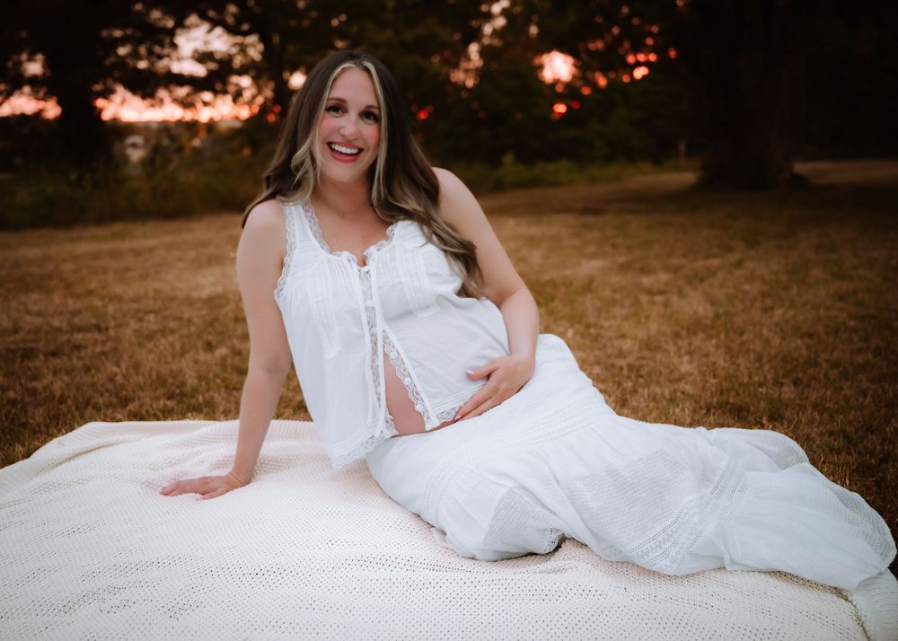 Pregnant woman in white dress sitting on blanket in golden hour field, cradling baby bump during maternity photo session.