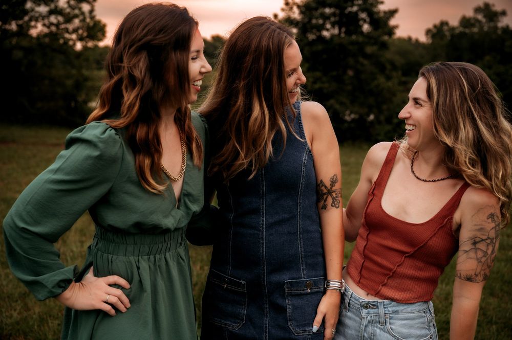 Three young women laughing and smiling together outdoors at sunset in a grassy field, wearing stylish casual outfits.