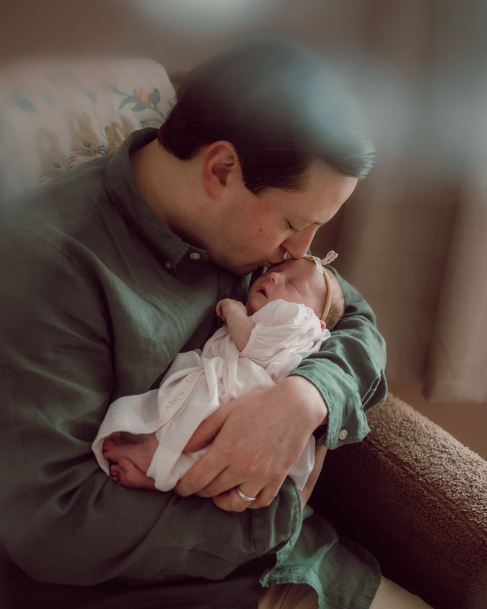 Parent in green shirt tenderly kissing newborn baby dressed in white, captured in warm indoor light.