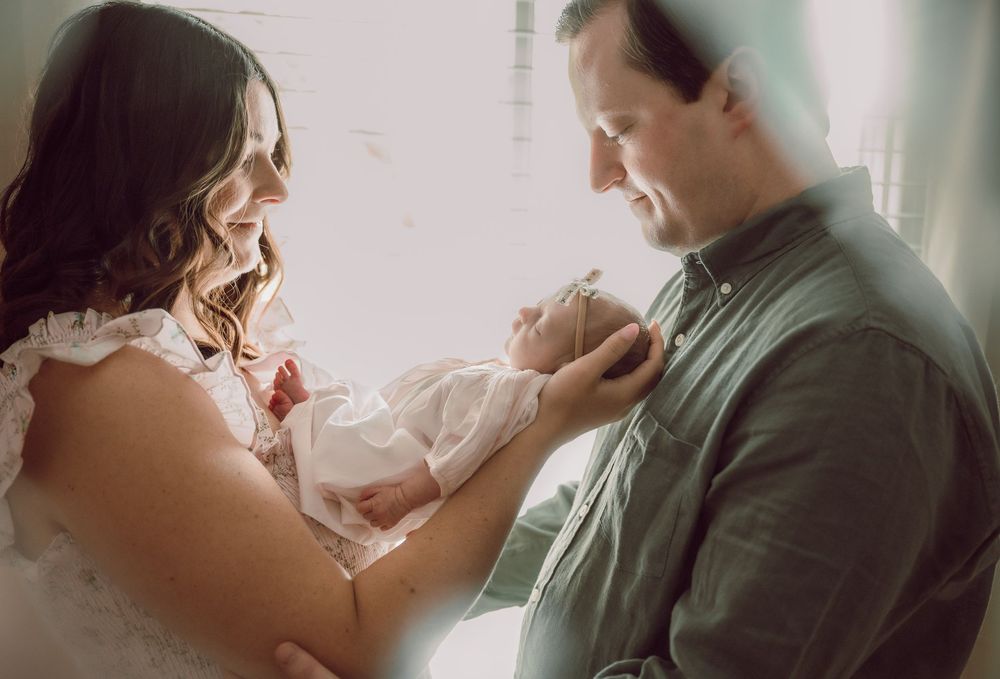New parents smiling lovingly at their newborn baby wrapped in white, sharing a tender family moment by a bright window.