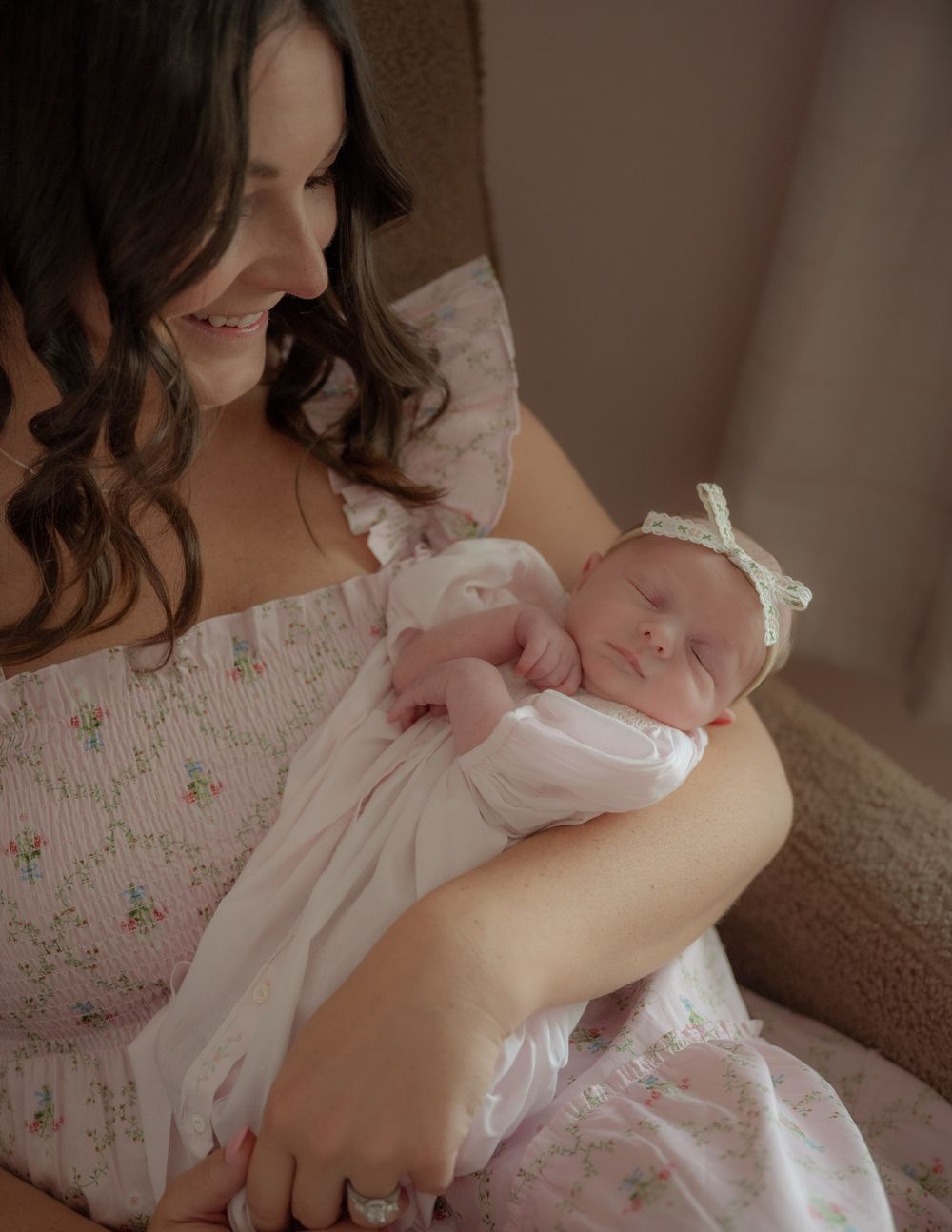A smiling mother with dark hair holds her sleeping newborn baby girl dressed in white with a bow headband.