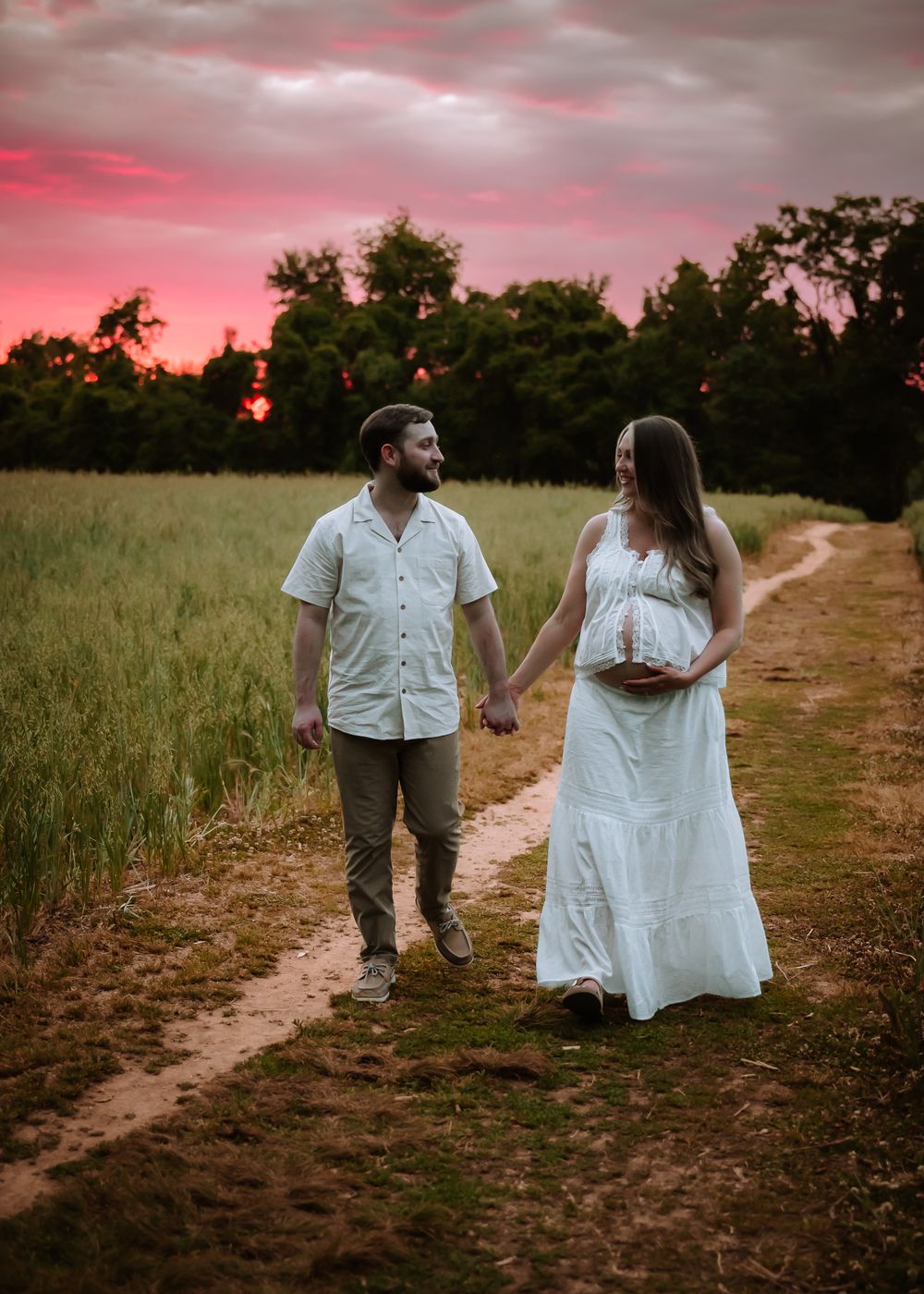 Couple holding hands on a dirt path at sunset, woman in white maternity dress cradling baby bump, vibrant pink sky backdrop.